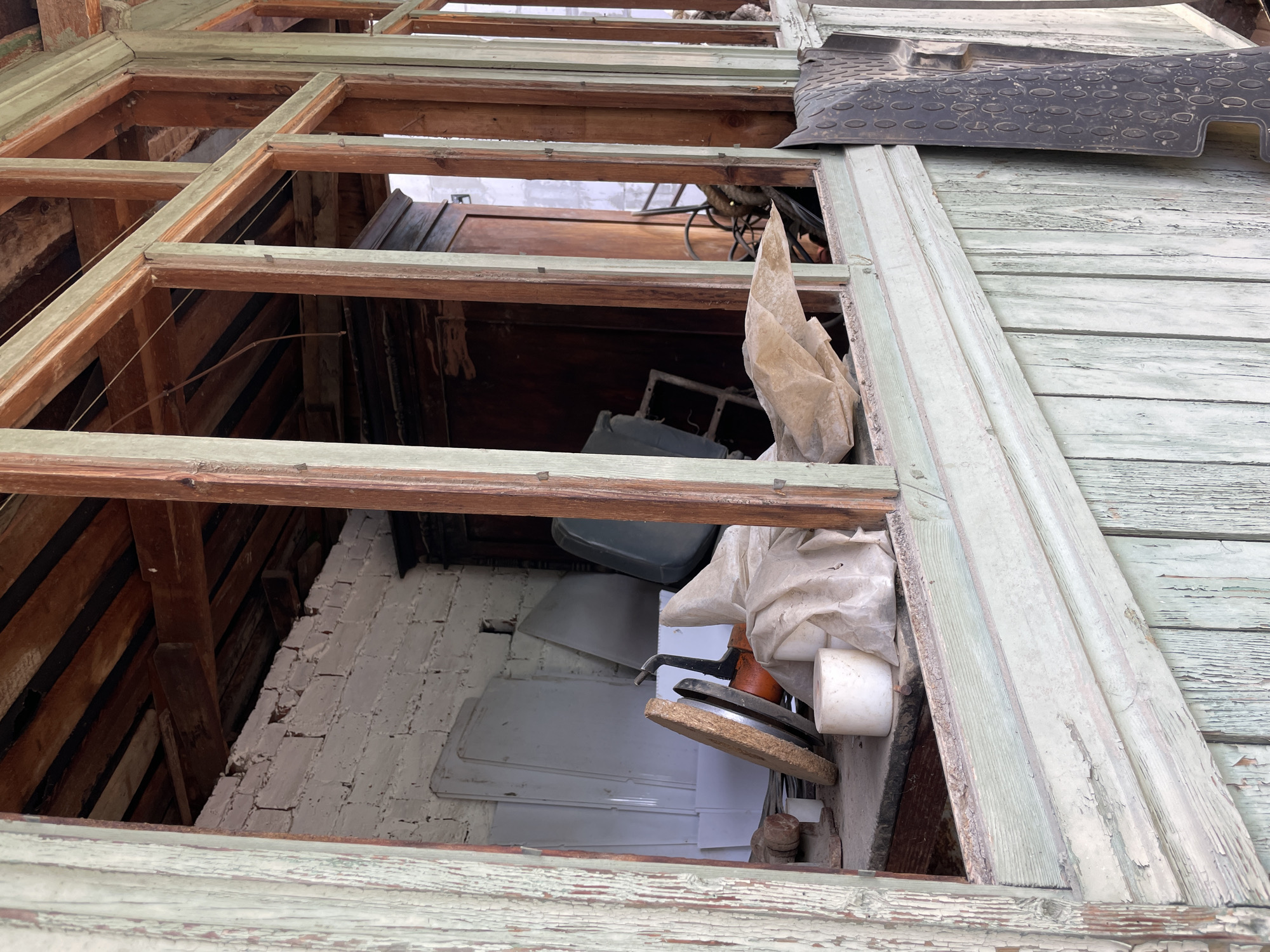 View looking through a deteriorated window frame, revealing the interior of a building. The window frame is made of wooden beams, some painted with light green paint that is peeling. Inside, a collection of objects are visible, including white sheets, a gray container, other miscellaneous items and a wooden object. The interior wall appears to be made of whitewashed brick. Outside the window, a section of weathered wooden plank siding is visible. A black rubber mat rests along the frame of the window opening. The location is Olginka, Russia. No people are visible. The image appears to be taken from a low vantage point looking up through the opening in the window.
