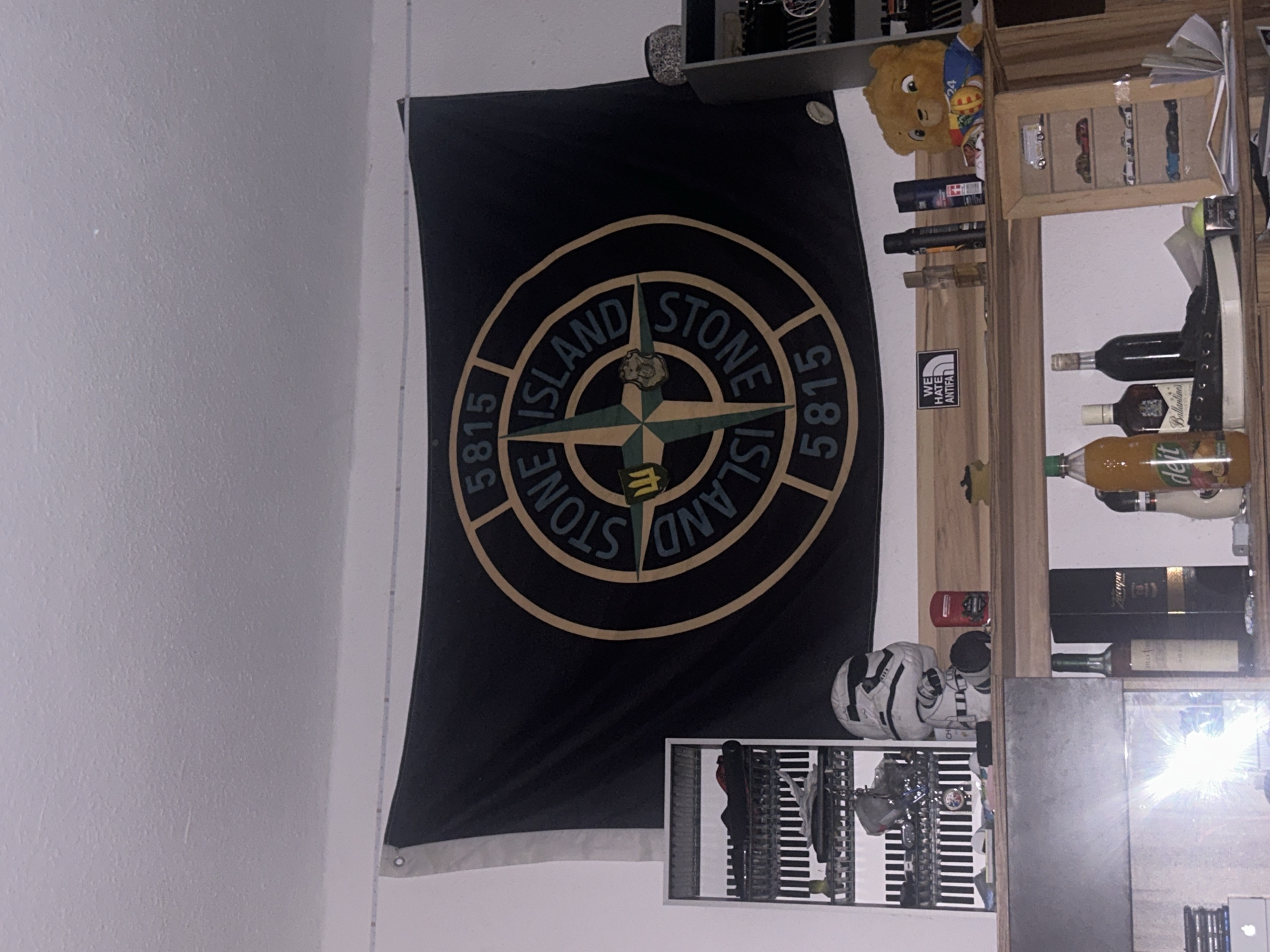 The image shows a room with a large black flag hanging on the wall. The flag has a circular emblem in the center with the words "STONE ISLAND" and numbers "5815" around it. To the left of the flag is a white shelf with various items, including shoes and what appears to be a Star Wars-themed figurine. Below the shelf, a portion of a metallic surface is visible, possibly a refrigerator or locker.
On a wooden shelf below the flag and the shelf unit, several items are displayed. These include bottles of liquor, a stuffed teddy bear, and other decorative objects. To the right, there is a dark cabinet or shelf unit containing more items.
The room appears to be a personal space, likely a bedroom or living area, decorated with personal items and memorabilia. The overall impression is of a lived-in space with a focus on specific interests, indicated by the flag and figurines. The lighting in the room suggests it is nighttime or the blinds are drawn, with artificial light sources illuminating the scene. The presence of a window with reflections of light suggests it is a room in a residential building. The information about the specific location in Gelsenkirchen, Germany is not present in the image itself.