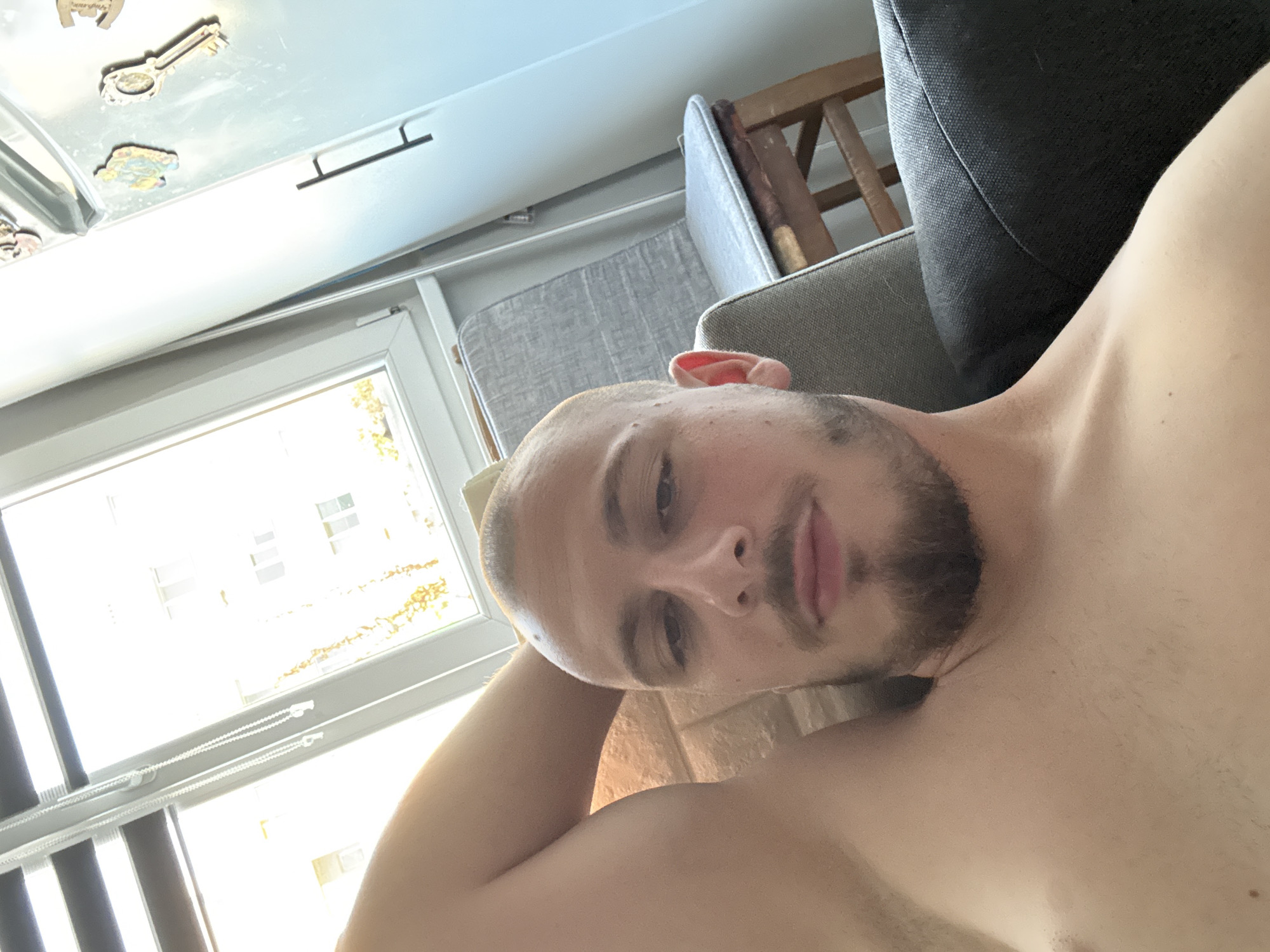 A shirtless man with a buzz cut and goatee reclines on a sofa, his right arm behind his head. Sunlight streams through a window behind him, illuminating a portion of the room. A gray upholstered chair with a wooden frame is visible to the man's right. The room appears to be an apartment in Stara Zagora, Bulgaria. A refrigerator with several magnets on its door is also in the background.