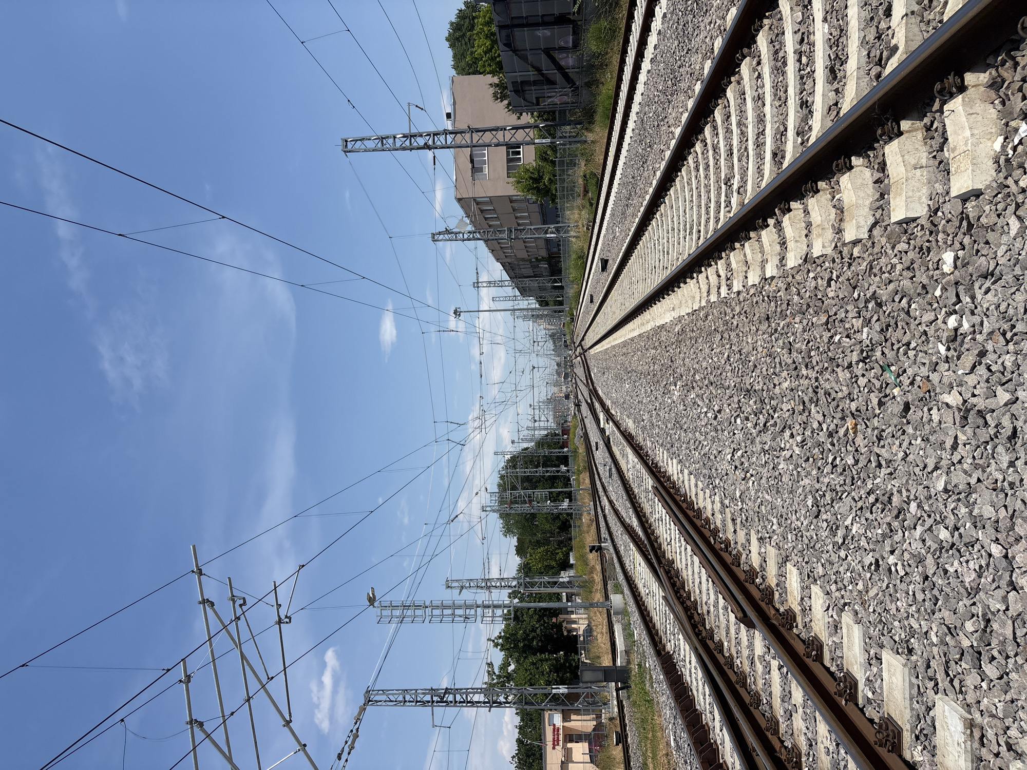 The image depicts a railway track with overhead power lines under a clear blue sky with scattered white clouds. The perspective is from ground level, looking down the tracks which stretch into the distance. The tracks themselves are made of metal rails supported by concrete sleepers and surrounded by gravel.
To the left of the tracks, a series of tall metal poles support a complex network of overhead wires, characteristic of an electrified railway system. Further back, a modern, multi-story building with a light brown facade and dark windows is visible, along with green trees and vegetation. The building appears to be an industrial or commercial facility.
The lighting suggests it is daytime, likely late morning or early afternoon, with bright sunlight casting shadows. The weather appears to be fair and sunny.
The scene is a typical urban or suburban railway environment, possibly in Plovdiv, Bulgaria, given the context. There are no people visible in the image, and the overall mood is one of functional infrastructure and quietness. A small red sign with white text is partially visible on a building in the distance, but the text is not legible. The image is oriented sideways, with the sky on the left and the ground on the right.