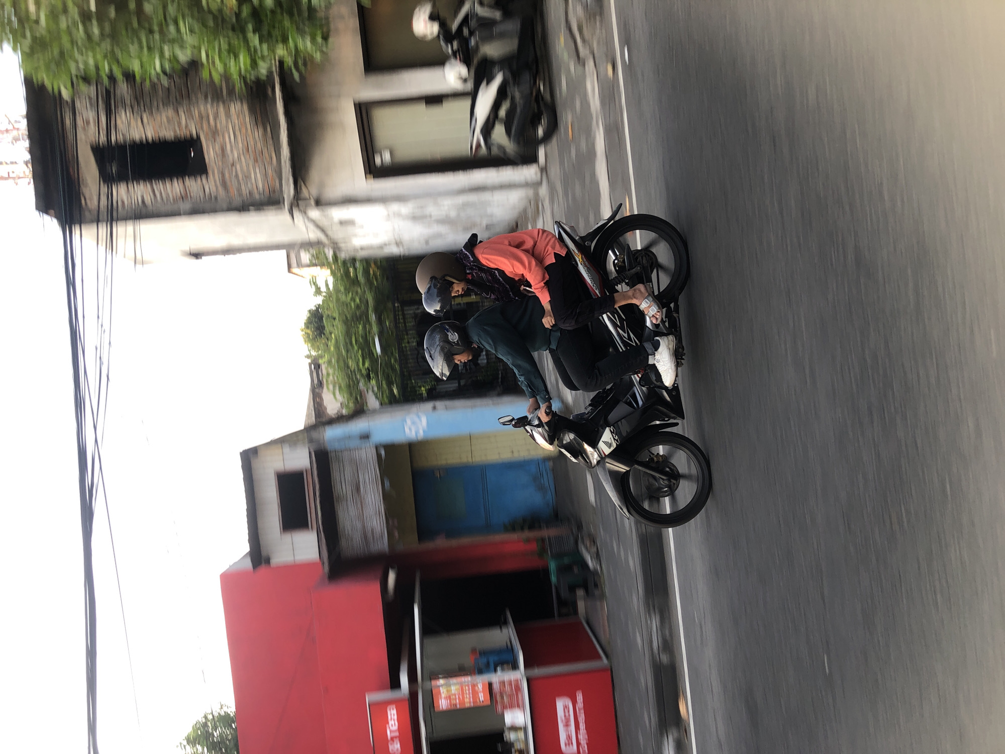 Two people are riding a black motorcycle down a street in Sidoarjo, Indonesia. The rider in front is wearing a dark green long-sleeved shirt and black pants, with a black helmet. The passenger behind them is wearing a bright orange long-sleeved shirt, black pants, and a patterned headscarf, with a light brown helmet. Both are wearing casual footwear. The motorcycle is in motion, indicated by the blur of the wheels and the background. To the left of the motorcycle, there is a red building with a sign that says "Teh" and another sign that says "Be Nice Coffee". Further down the street are buildings with various architectural styles, including some that appear older and in disrepair, and trees. The sky is overcast. There is another motorcycle parked in the background to the right.