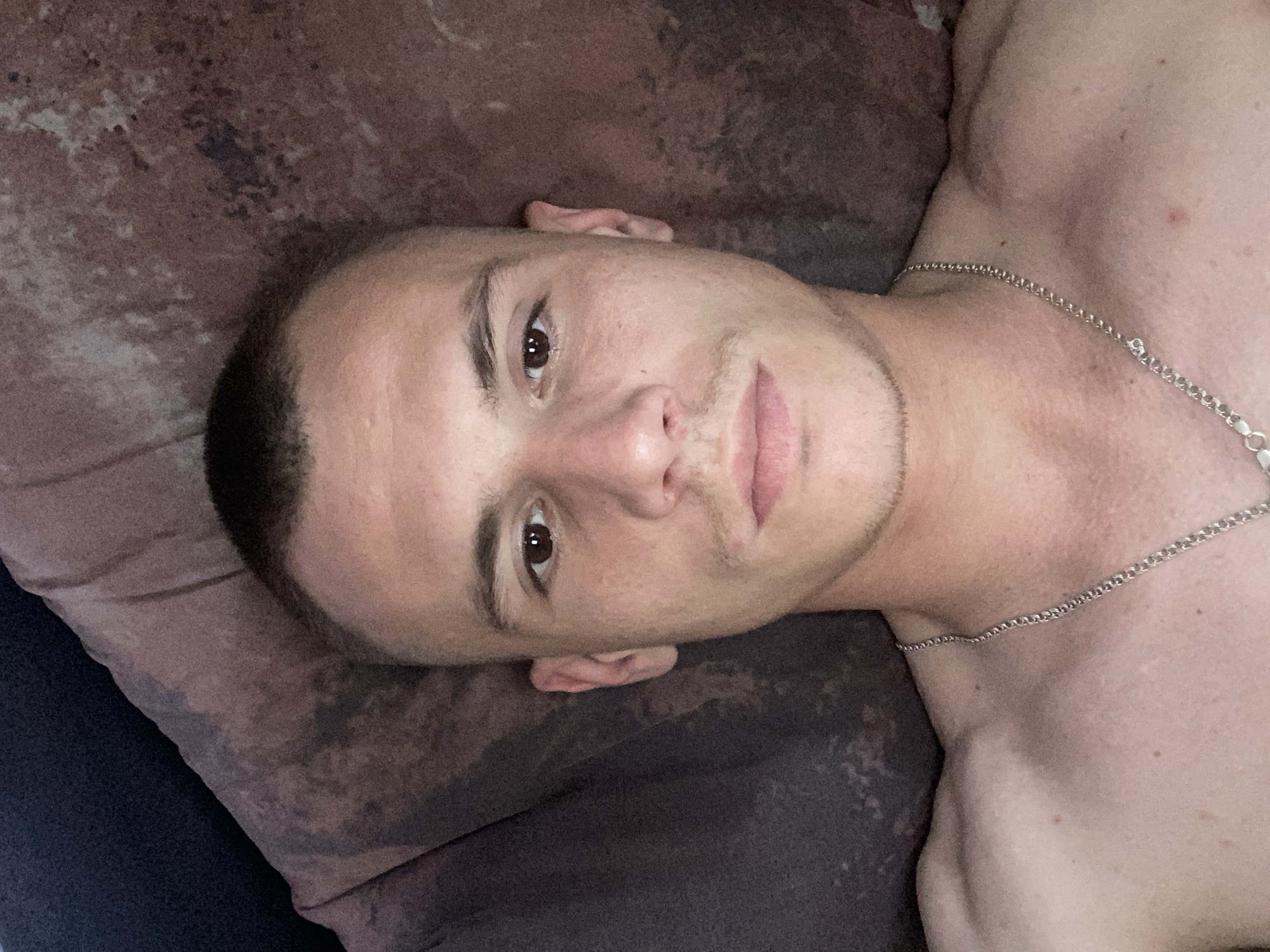 The media file is a close-up, low-angle, horizontal shot of a young man lying on his side on a brown surface, possibly a bed or couch. The man has short, dark hair, brown eyes, and fair skin with some blemishes. He is wearing a silver chain around his neck. His face is partially obscured by the angle, but his left eye, nose, and mouth are visible. The background is a textured, dark brown fabric, and a darker blue fabric is visible on the left edge of the frame. The lighting appears to be dim and indoor, suggesting it might be nighttime or a dimly lit room. There is no discernible activity or event taking place; the man appears to be resting or posing for the camera. The overall mood is relaxed and intimate due to the close-up nature of the shot and the subject's relaxed posture. No text is visible in the image.