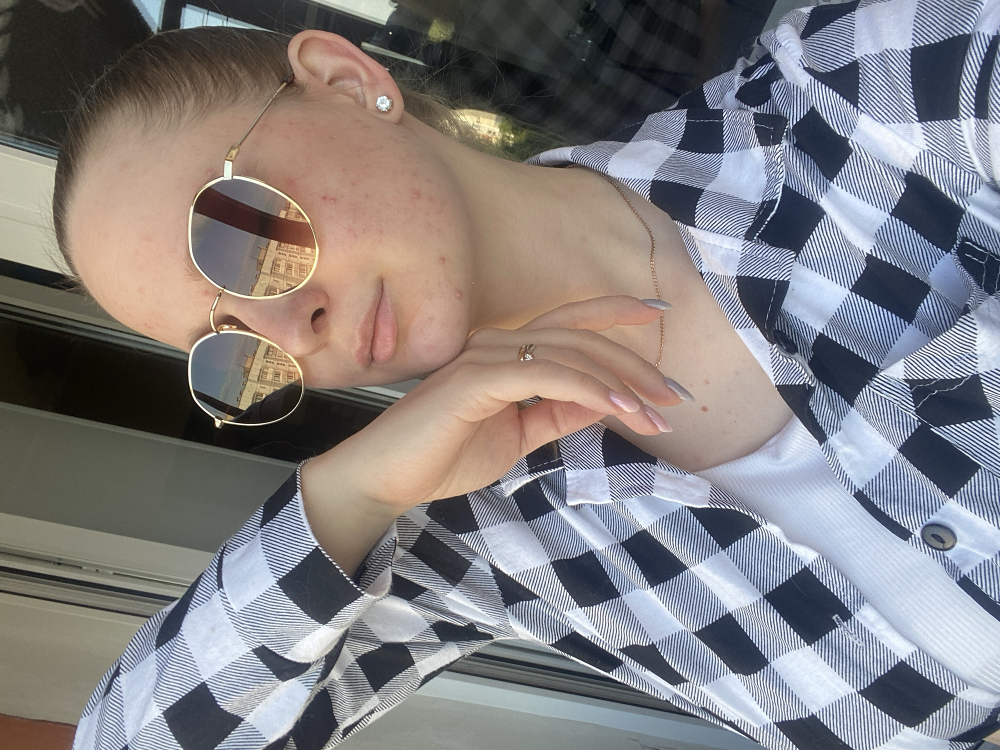 A young woman with blonde hair pulled back is captured in a close-up, side-profile selfie, leaning against a window frame. She wears round, gold-rimmed sunglasses that reflect buildings and a clear sky. Her left hand, adorned with a ring, rests gently on her chin, and her nails are painted in a light gray hue. She sports a black and white checkered shirt open over a white ribbed tank top. A delicate gold necklace is visible around her neck. The background suggests an urban setting with architectural elements visible through the window. The lighting indicates it is daytime.