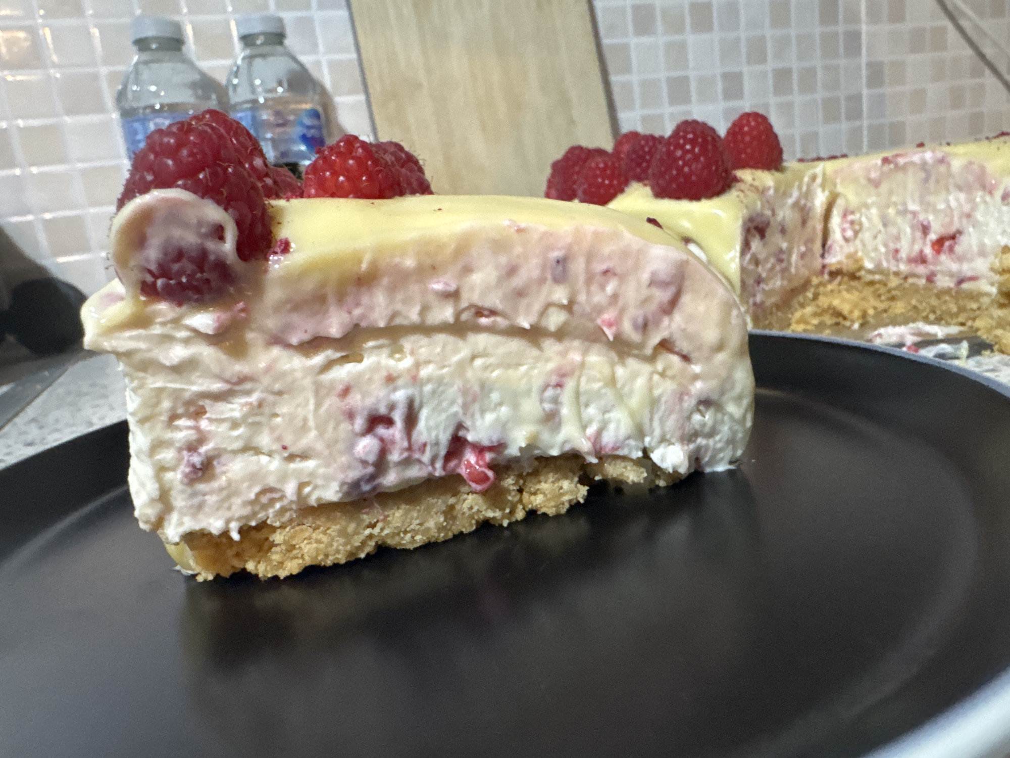 A close-up shot depicts a slice of raspberry cheesecake on a black plate. The cheesecake has a crumbly crust, a creamy filling with visible raspberry pieces, and a yellow topping adorned with fresh raspberries. Additional bottles are behind it. The scene is set indoors, with a tiled backsplash in the background. Given the request, the location is presumed to be in Desborough, United Kingdom, although this cannot be determined solely from visual information.