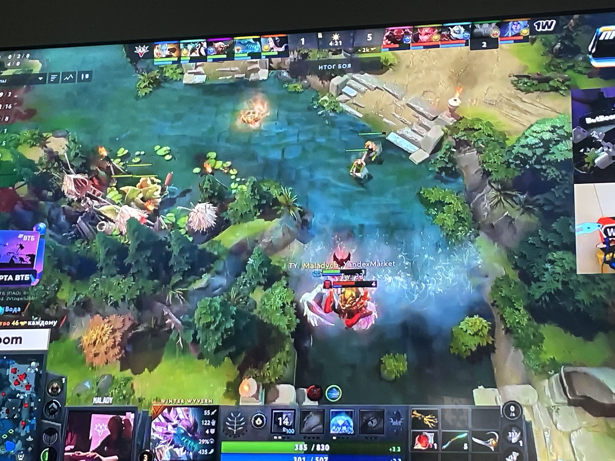 The image shows a screenshot of a video game, likely Dota 2, displayed on a computer monitor. The game features a top-down perspective of a fantasy landscape with a body of water, trees, and terrain. Several in-game elements are visible, including player avatars, health bars, a minimap in the bottom left corner, and various interface elements displaying statistics, item icons, and text. One player is identified by the name "TY, Maladych, YandexMarket" and is engaged in combat with another player. The game's interface indicates a battle is in progress, with scores and timers visible. There are no discernible real-world locations or people present within the image itself, only the digital representation within the video game. Therefore, the location in the city of Sokol'ya Griva, Russia, is not depicted or suggested by the visual content.
