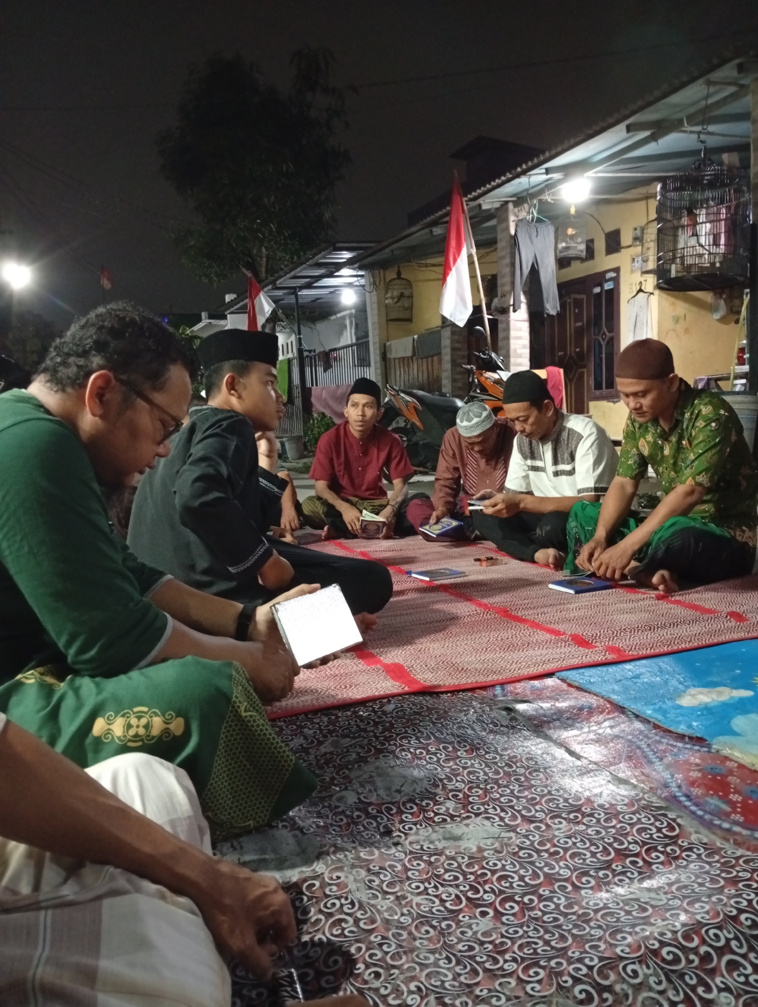 In the city of Caringin, Indonesia, at night, a group of Muslim men are gathered on mats on the ground outside a building. They are all wearing traditional Islamic attire, including peci caps and long-sleeved shirts or robes. Most of the men are looking down, either at books or at something in their hands, suggesting they are engaged in religious study or prayer. One man on the far left is wearing glasses and looking down at a white rectangular object, possibly a tablet or a book. Another man in the center is holding a book. In the background, there is a building with a corrugated metal roof, a flag of Indonesia, and a birdcage. The overall scene depicts a communal gathering for religious purposes.