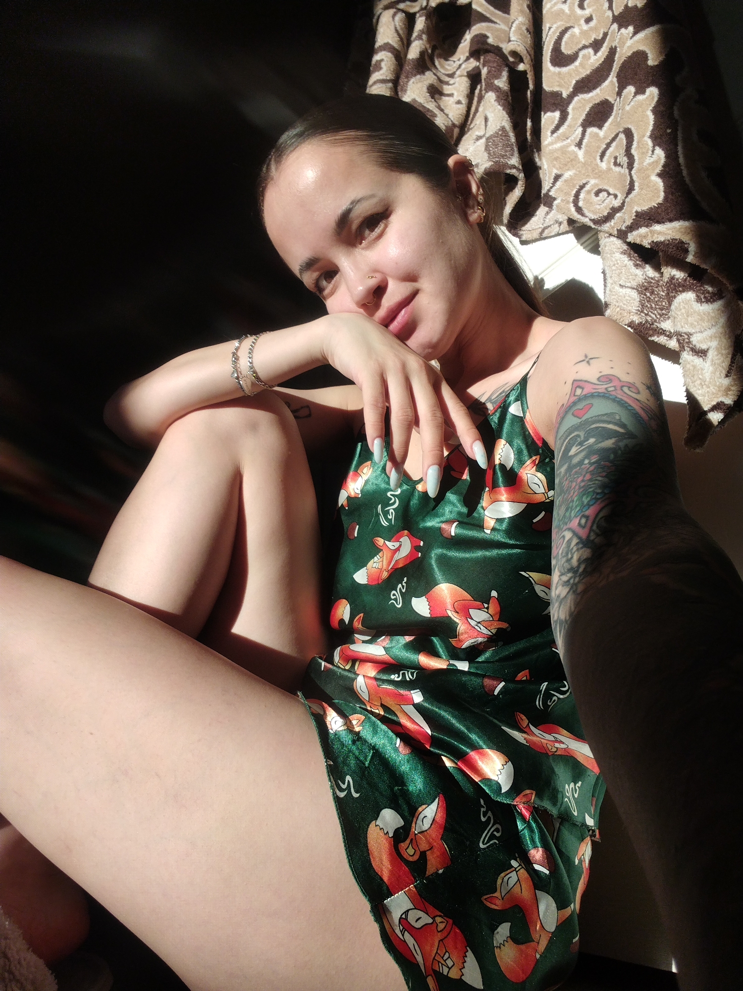 A woman with dark hair pulled back, wearing a green satin pajama romper with an orange fox print, is posed indoors in a spot with harsh lighting. She is resting her left arm on her bent left leg, with her hand near her face. Her fingernails are long and painted a light color. She has multiple piercings in her left ear and a nose ring. A silver bracelet is visible on her left wrist. Her right arm, heavily tattooed, extends toward the right edge of the frame. A patterned brown and beige textile is visible in the background. The specific location within the city of Unknown, Unknown is undeterminable.