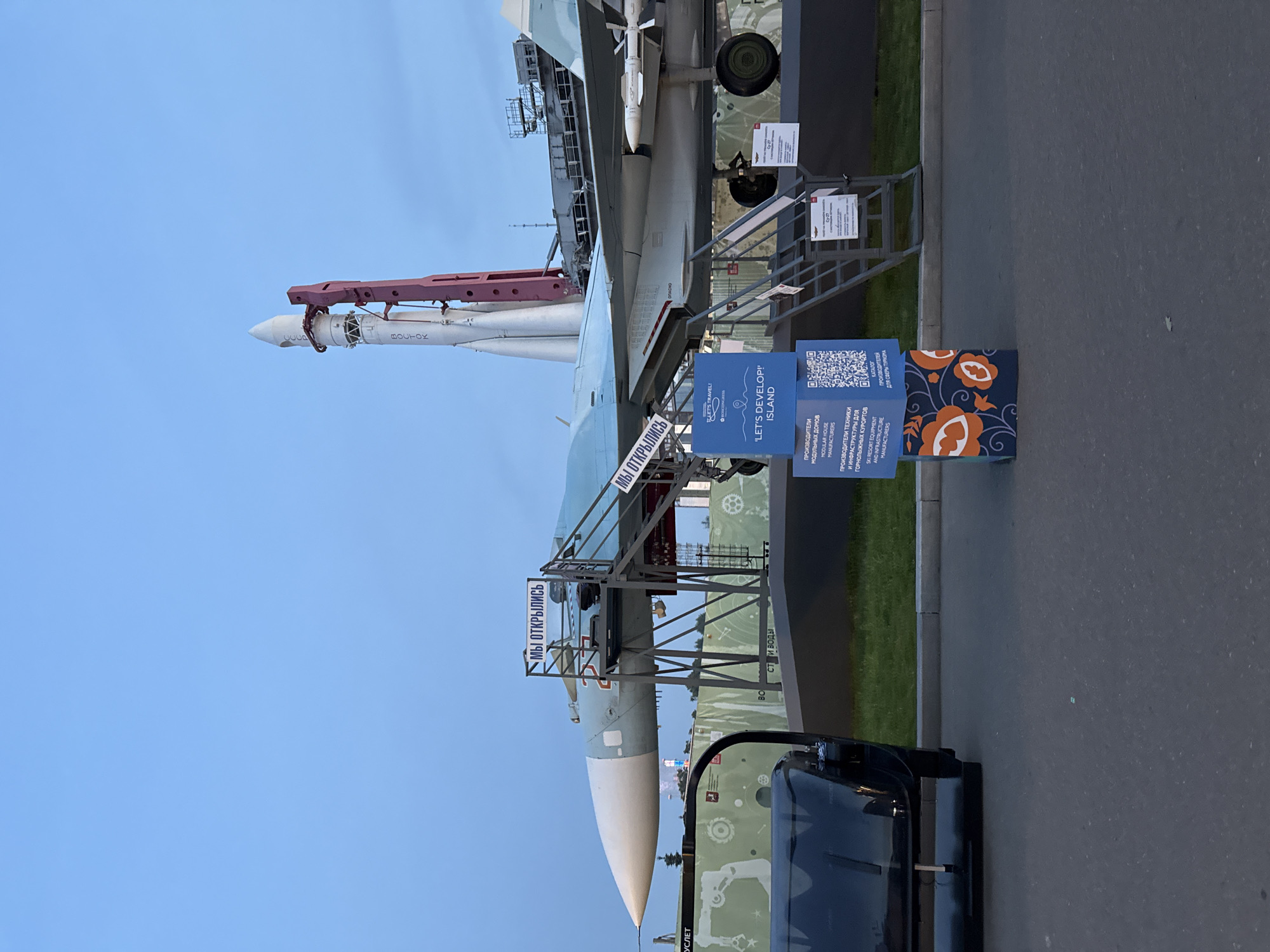 The image depicts an outdoor exhibition scene in Moscow, Russia, featuring a fighter jet and a rocket on display. The fighter jet, a Sukhoi Su-27, is parked horizontally on a raised platform, with a missile mounted on its wing. Stairs lead up to the jet for visitors to access it. Behind the jet, a white and red rocket stands vertically, suggesting a space or aviation-themed exhibition. In the foreground, a portion of a dark vehicle is visible on the left. Several blue informational stands are placed in front of the exhibition area, one of which features a QR code and text in Russian and English. The sky is a clear, pale blue, indicating it is likely late afternoon or early evening. The ground appears to be asphalt and grass. The overall setting suggests a public event or museum exhibit focused on aerospace technology.
