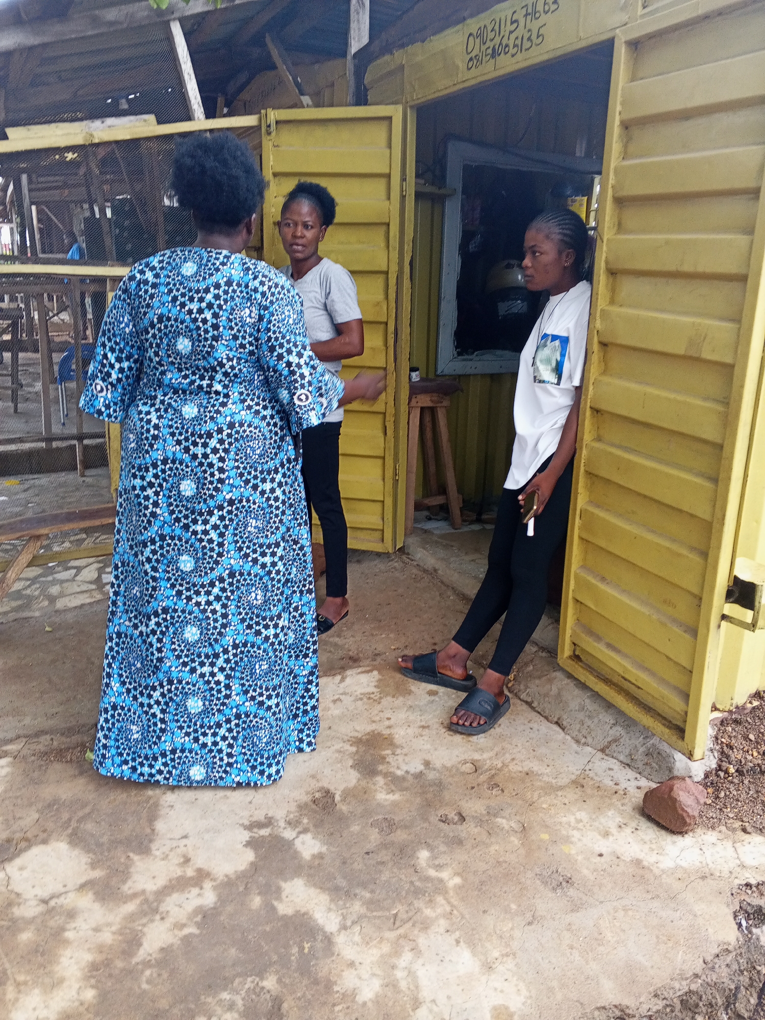 The image shows three women in what appears to be Zango-ndaji, Nigeria. One woman, seen from the back, wears a long, blue patterned dress. Another woman in a gray shirt and black pants stands nearby. A third woman leans against the doorway of a yellow building, wearing a white t-shirt and black leggings. The yellow building has a phone number (090311571663/08156065135) printed above the doorway. There is a stool inside the building near the window. The foreground is a concrete area with a rock near the building. An open-air structure with a table and chairs is visible in the background.