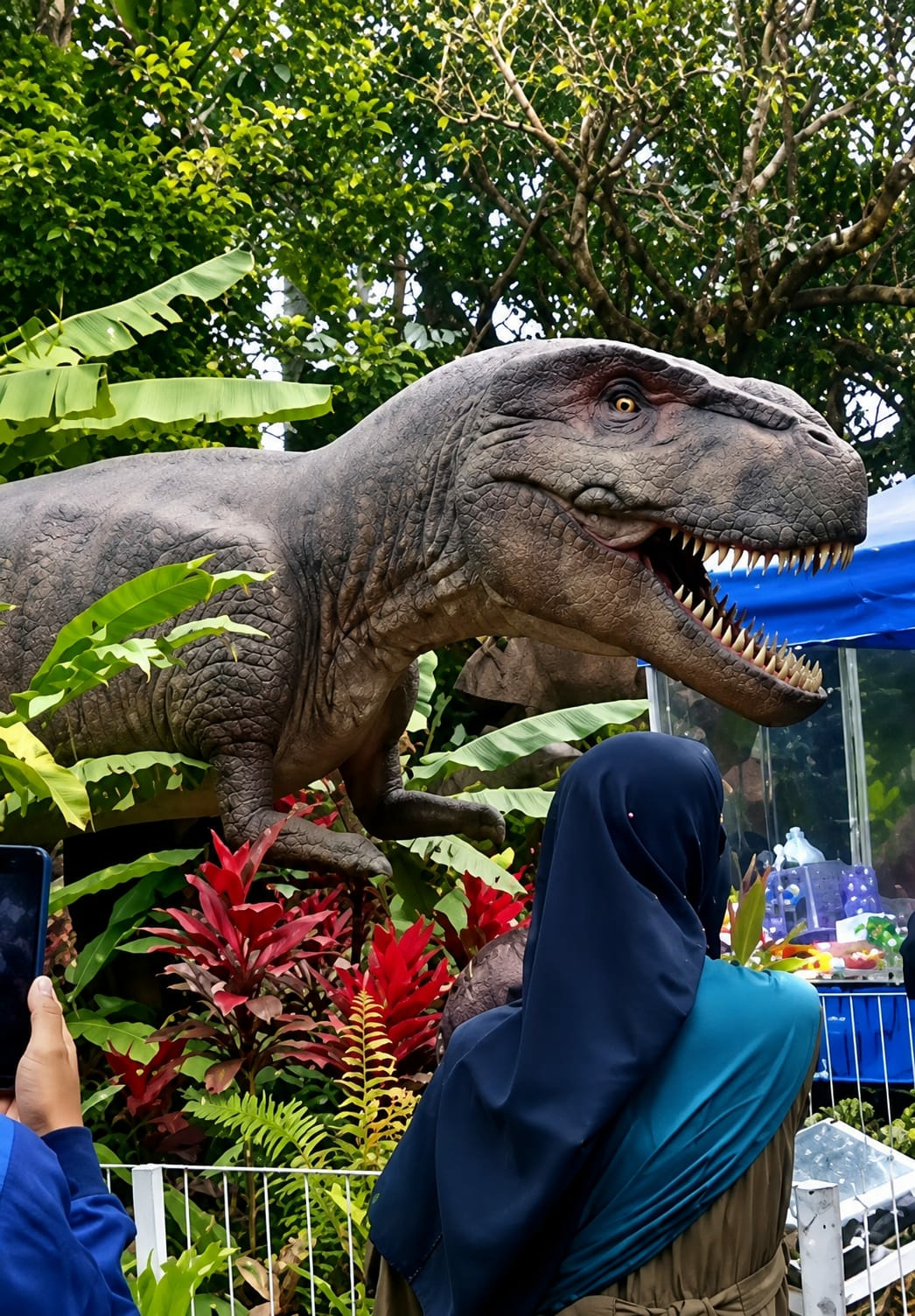 A realistic dinosaur statue, resembling a Tyrannosaurus Rex with its mouth open and sharp teeth, is positioned amidst lush greenery and red plants. A person wearing a dark blue hijab and a greenish-blue shawl is standing in front of the dinosaur, facing away from the camera, and holding a smartphone to take a picture. Another person's arm, wearing a blue jacket and holding a black smartphone, is visible on the left side of the frame. In the background, a blue tarp covers a structure, and beyond that, some glass displays with colorful items are visible. The overall scene appears to be an outdoor exhibit, possibly in a park or theme attraction, located in Bandar Baru, Indonesia, judging by the surrounding tropical vegetation.
