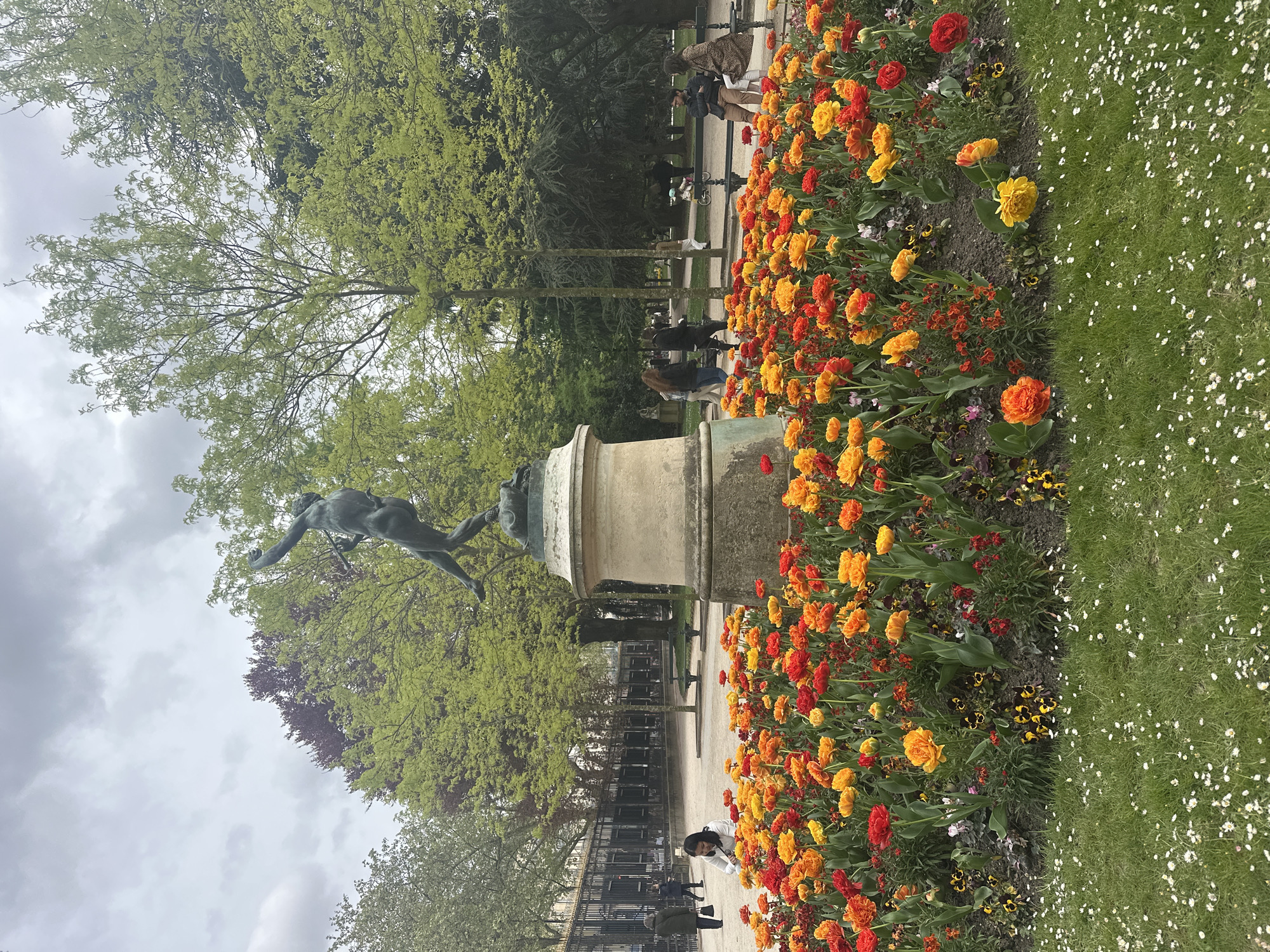 The image captures a scene in a Parisian park, featuring a statue of a nude figure in motion atop a stone pedestal. The statue is surrounded by vibrant flower beds, predominantly filled with orange, yellow, and red tulips. Lush green grass borders the flower beds. In the background, a wrought iron fence separates the park from a paved walkway where people are strolling. Tall trees with green foliage are visible behind the fence, partly obscuring the sky, which appears overcast. The photograph is taken at ground level, with a slightly low angle capturing the statue and the flowers.