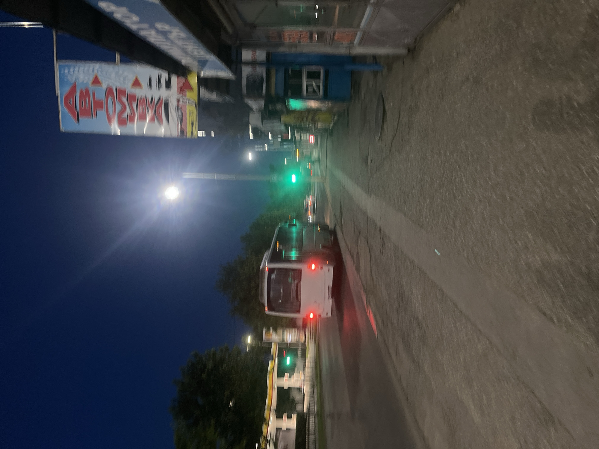 The image depicts a street scene at dusk or night in Plovdiv, Bulgaria. A white bus is visible on the right side of the road, moving away from the viewer, with its taillights illuminated red. The bus has several green lights visible through its windows on the right side. A gas station with green traffic lights is situated on the left side of the street. A tall vertical sign with red and white lettering, possibly advertising an auto service, is prominent in the background on the right. Buildings line the right side of the street, featuring storefronts and awnings. The street is paved with asphalt and sidewalks are present. The sky is dark blue, indicating a nighttime setting, and a streetlamp with a bright light source is visible in the upper center of the frame, casting a glare. Trees are present on the far left of the image.