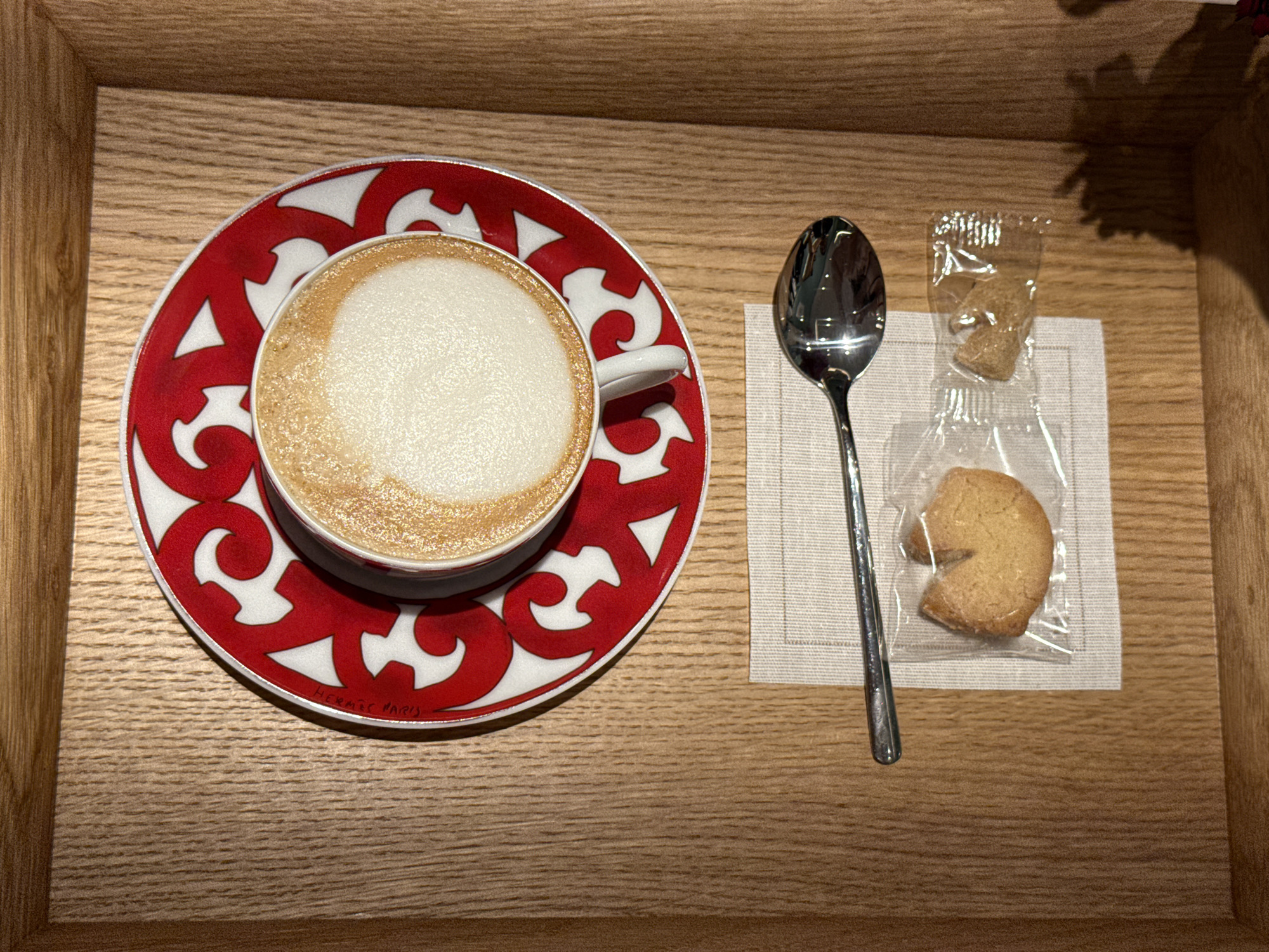 The image shows a latte with a red and white patterned saucer, a spoon, a small cookie in a plastic bag, and a piece of brown sugar in a plastic bag, all resting on a beige napkin within a light brown wooden tray. The saucer bears the inscription "HERMÈS PARIS" around its edge. The wooden tray has a distinct wood grain pattern. Based on available information, this scene is likely from somewhere in Zapopan, Mexico, but the image itself does not offer any specific clues to its exact location.