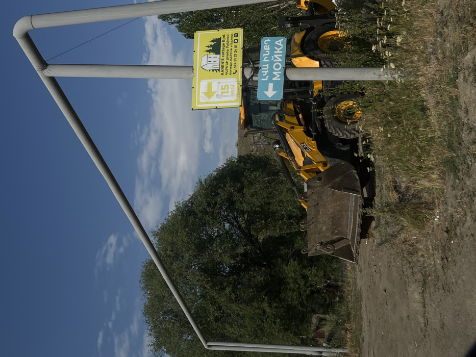 A yellow JCB backhoe loader sits beside a dirt road under a partly cloudy sky. Two signs are fixed to a metal frame. The first sign advertises "KAREN'S cottage Bjni" with information in Armenian, English, and Russian, including a phone number. The second sign, with a blue arrow, points to "ԼՎԱ ԻՆՔԴ МОЙКА," which translates to "Wash It Yourself Car Wash." A 15 km/u speed limit sign is also present. Green trees line the area.