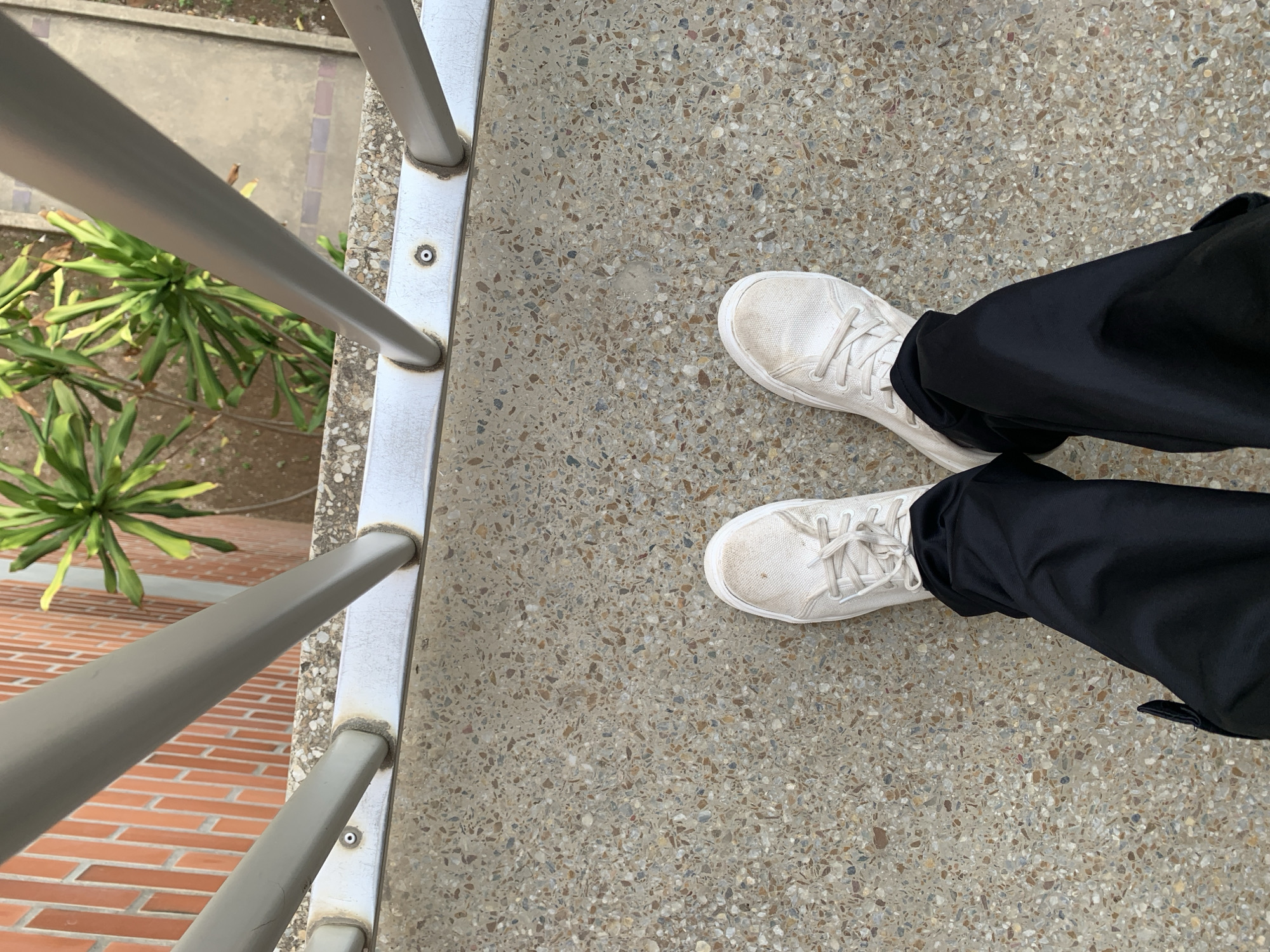 A first-person perspective shot shows a person wearing black pants and dirty white sneakers standing on a concrete surface with embedded pebbles. A metal railing with gray posts runs along the top of the image, with a brick wall and some greenery visible behind it. The scene appears to be outdoors, possibly on a balcony or walkway, with no specific identifying features of Las Piedras, Venezuela.