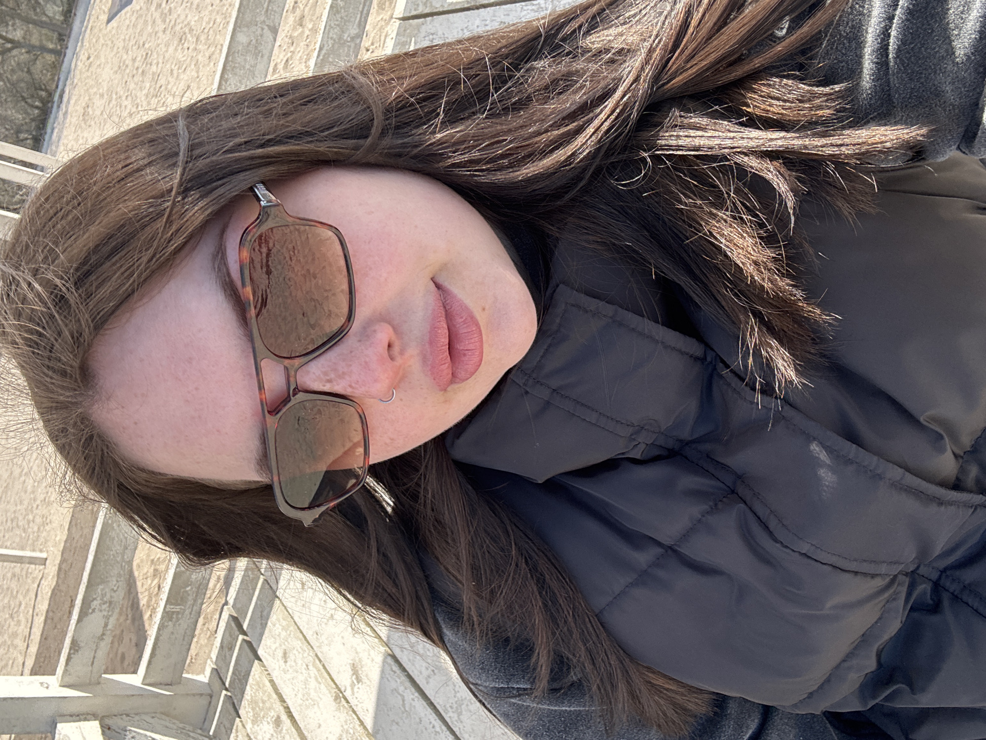 A young woman is posing for a selfie. She is wearing sunglasses and a black puffer jacket. She has dark brown hair. The location is Chornomorskoe, Ukraine. The background appears to be a light-colored, possibly concrete or stone, outdoor structure.