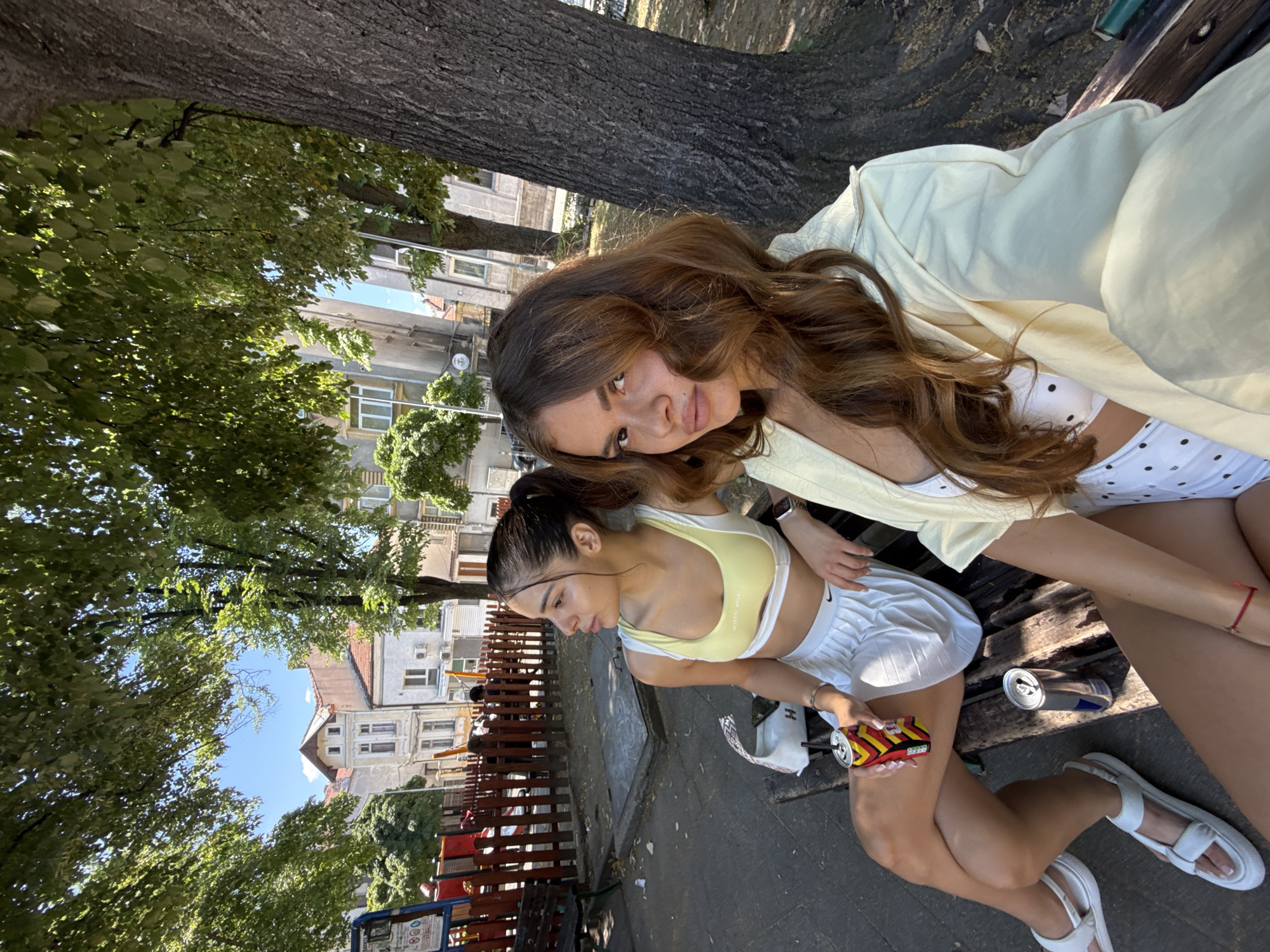 Two young women are sitting on a wooden park bench in Plovdiv, Bulgaria. The woman on the left is wearing a yellow sports bra and a white pleated skirt. She is holding a can of soda with a red, yellow, and black design and has a white bag next to her. The woman on the right is wearing a white bikini top with black polka dots and a light yellow jacket. She has long brown hair and is looking at the camera. In the background, there is a playground with a red fence, trees, and apartment buildings. A large tree trunk is visible on the right side of the image. The ground is paved, and there are fallen leaves scattered around. The scene appears to be set on a sunny day.