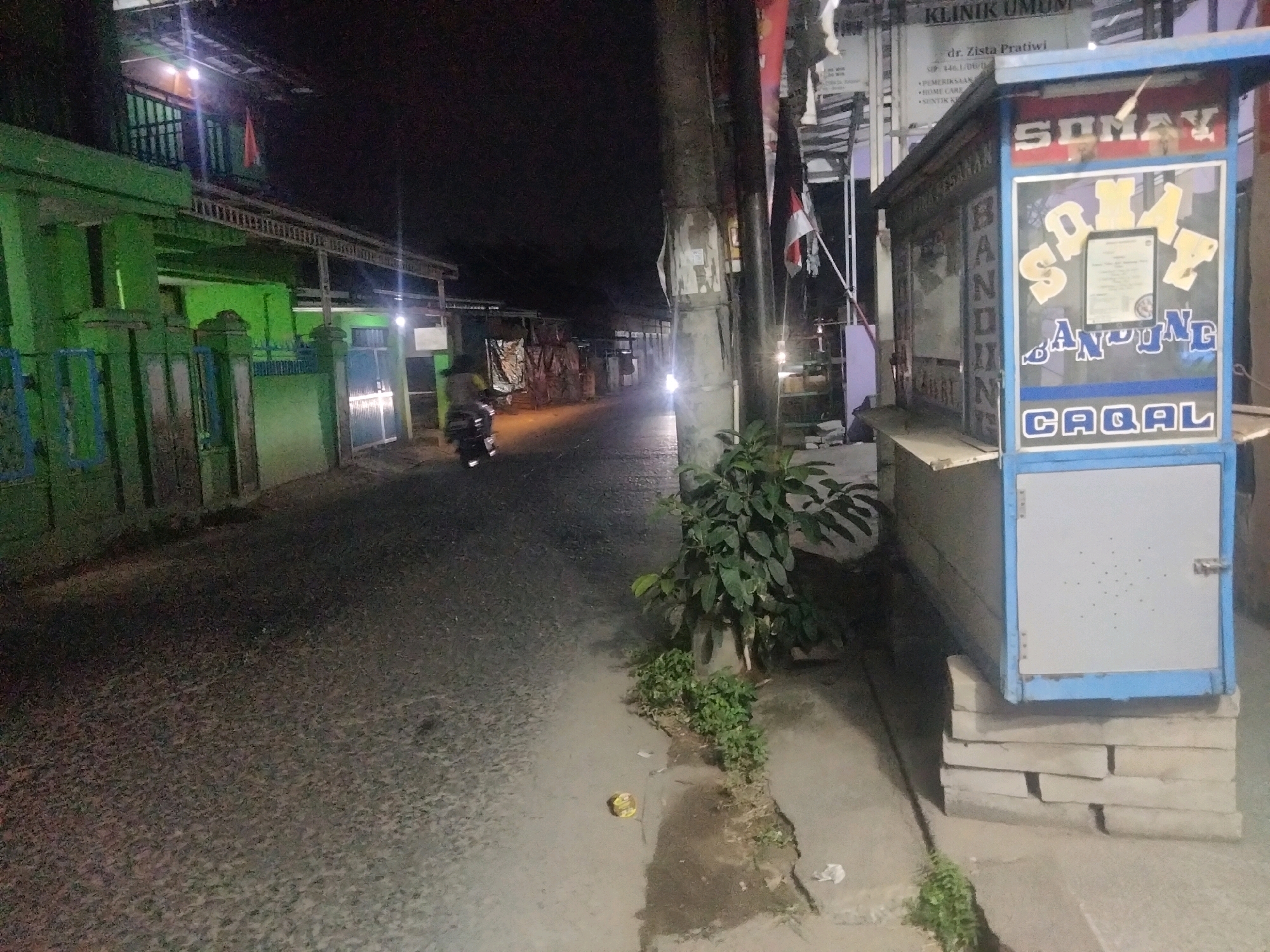 The image captures a dimly lit street scene at night in Suka Sari, Indonesia, likely a residential area or a small commercial strip. On the right side of the frame, a food stall is prominently displayed, painted in blue and white. The stall features lettering that reads "SOMAY" in yellow, with "BANDUNG" in blue lettering below it. The word "CAQAL" is also visible at the bottom of the stall. A small, framed document or certificate is attached to the stall, positioned above the "BANDUNG" text. To the left of the stall, a young plant is growing near a utility pole.
The street itself appears to be paved with asphalt and shows signs of wear. In the distance, a motorcycle with its headlights on is traveling away from the camera, illuminating a section of the road. On the left side of the street, there are buildings with green facades and metal fencing. The overall atmosphere is quiet and somewhat subdued due to the nighttime setting and the limited illumination. The presence of the food stall suggests a local vendor operating in the area, catering to the community. The background is largely obscured by darkness, with hints of other structures and foliage.