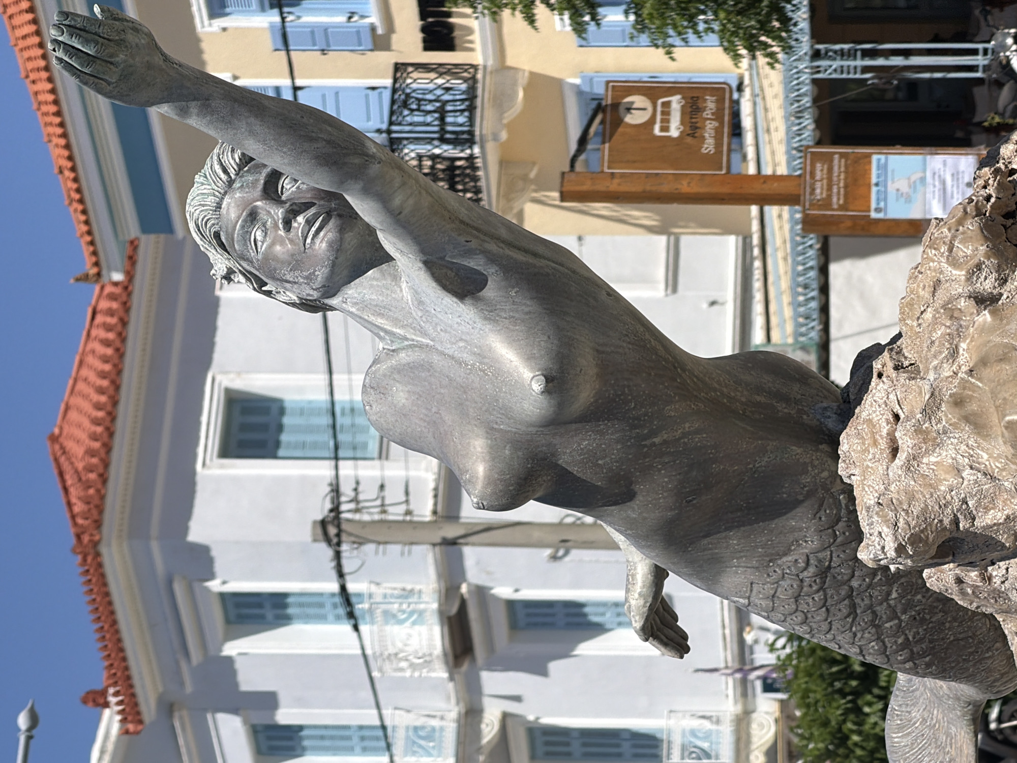 A medium close-up shows a bronze statue of a mermaid against a background that includes a building, blue sky, and signs, located in the city of Poros, Greece. The statue is in the foreground. The mermaid is depicted with a human upper body and a fish tail, attached to stone that makes up its base. The mermaid is looking upward and slightly to the left. Her left arm is raised.
The background includes buildings, with blue window shutters. Above the mermaid’s back are two wood signs that are mounted on a single post. The first sign says, “Αφετηρία Starting Point” and depicts a small train with seats in it. The second sign below, which says “Tourist Information,” shows a small map.