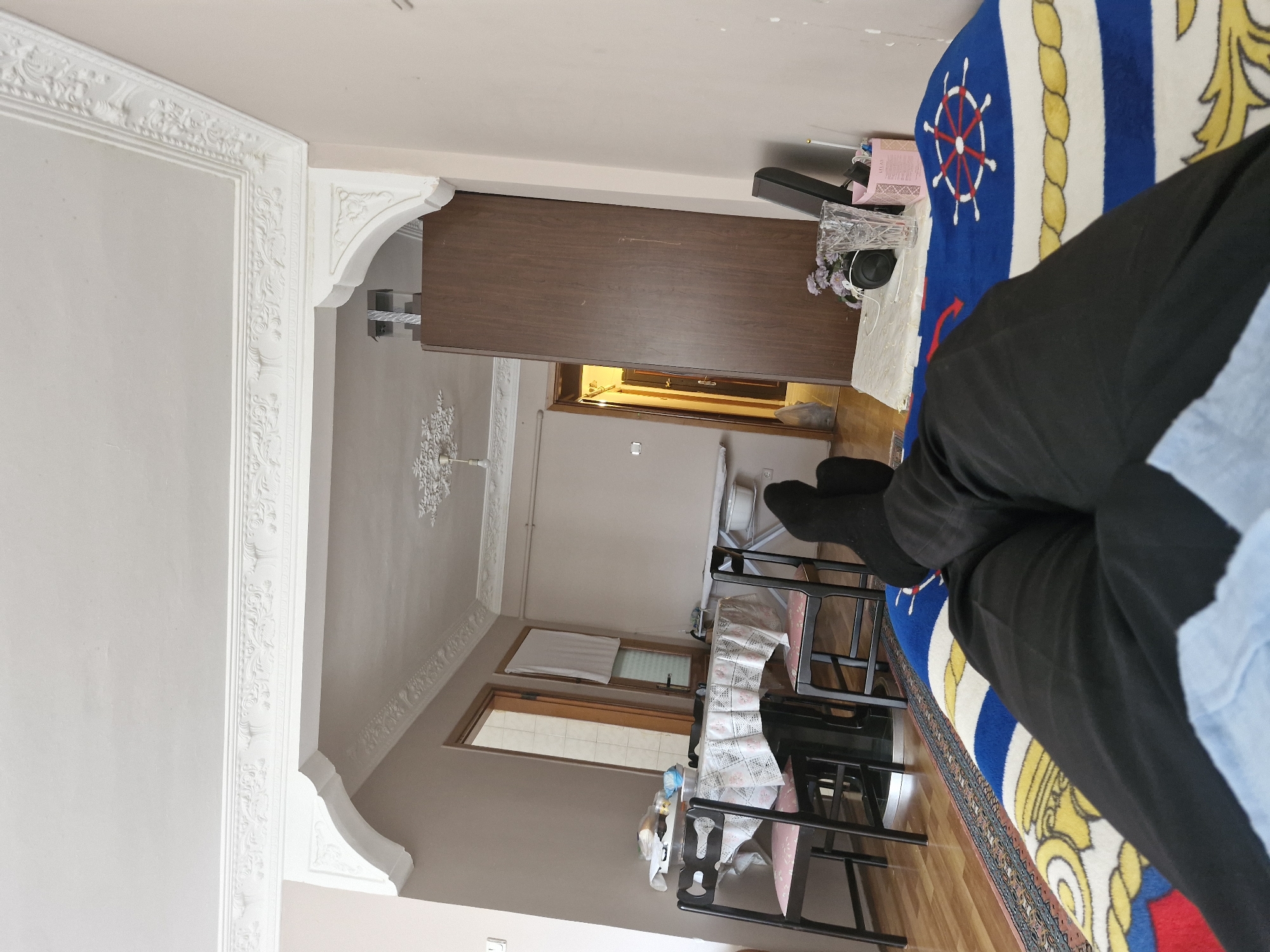 The image depicts an indoor scene, likely a living room or dining area, from a first-person perspective. The foreground shows a person's legs crossed, clad in black pants and black socks, resting on a blanket with a blue and white striped pattern featuring a ship's wheel and gold rope motif.
The background reveals a dining table with four chairs. The table is covered with a patterned tablecloth. On the table, there are various items including a white container, possibly food packaging, and a blue plastic bag. To the left of the table, there is a closed door and a window covered with a white curtain.
Further into the background, a doorway leads to another room where a hallway is visible, with another door at the end. To the right of the dining area, a tall, dark brown wooden wardrobe stands against a pale pink wall. On top of a surface next to the wardrobe, there is a speaker, a crystal vase with a small bouquet of flowers, and a pink container.
The ceiling features intricate white decorative molding, suggesting an older or elaborately decorated building. An archway with similar decorative molding frames the view into the dining area. The overall lighting appears to be artificial.
While the image is not geotagged, the style of the interior decoration, particularly the ceiling molding and the type of furniture, is consistent with residential interiors found in Turkey, including areas like Küçükçekmece.
