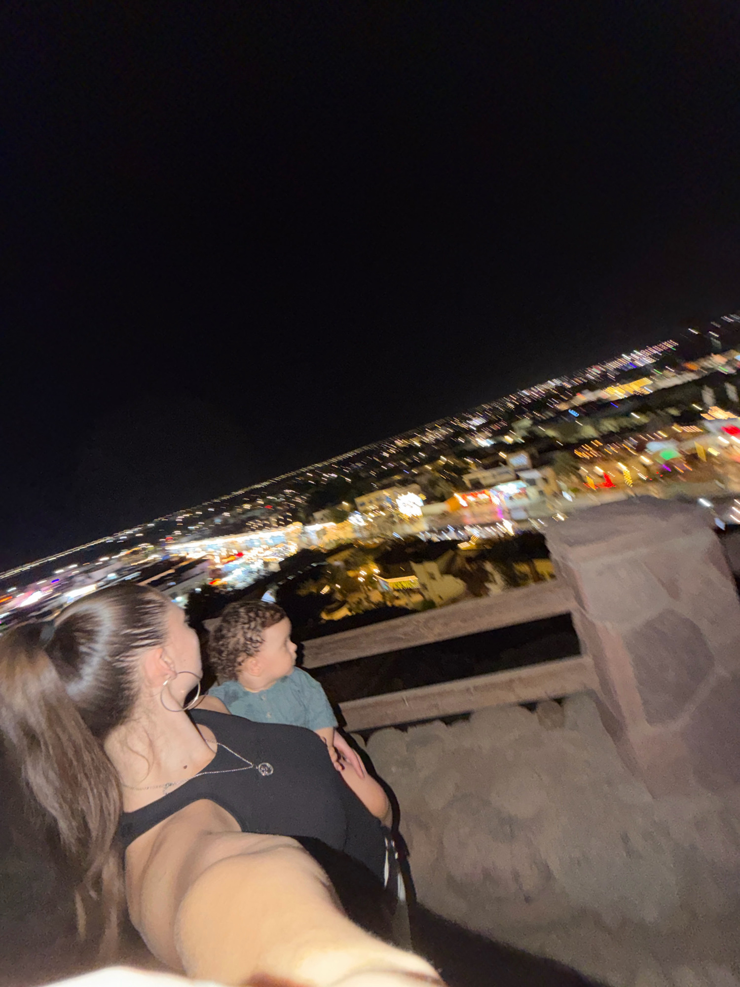 A woman with long, dark hair pulled into a ponytail and a silver hoop earring is looking out over a brightly lit city at night, with a young child in a blue shirt standing next to her, also looking out. The woman is wearing a black top and a silver necklace. They are standing on a stone ledge with a wooden railing in front of them. The city lights stretch out into the distance, illuminating buildings and streets. The sky is dark and clear. The image appears to be taken in Sharm El Sheikh, Egypt, possibly from a vantage point overlooking the Naama Bay area, known for its vibrant nightlife and illuminated landscape.