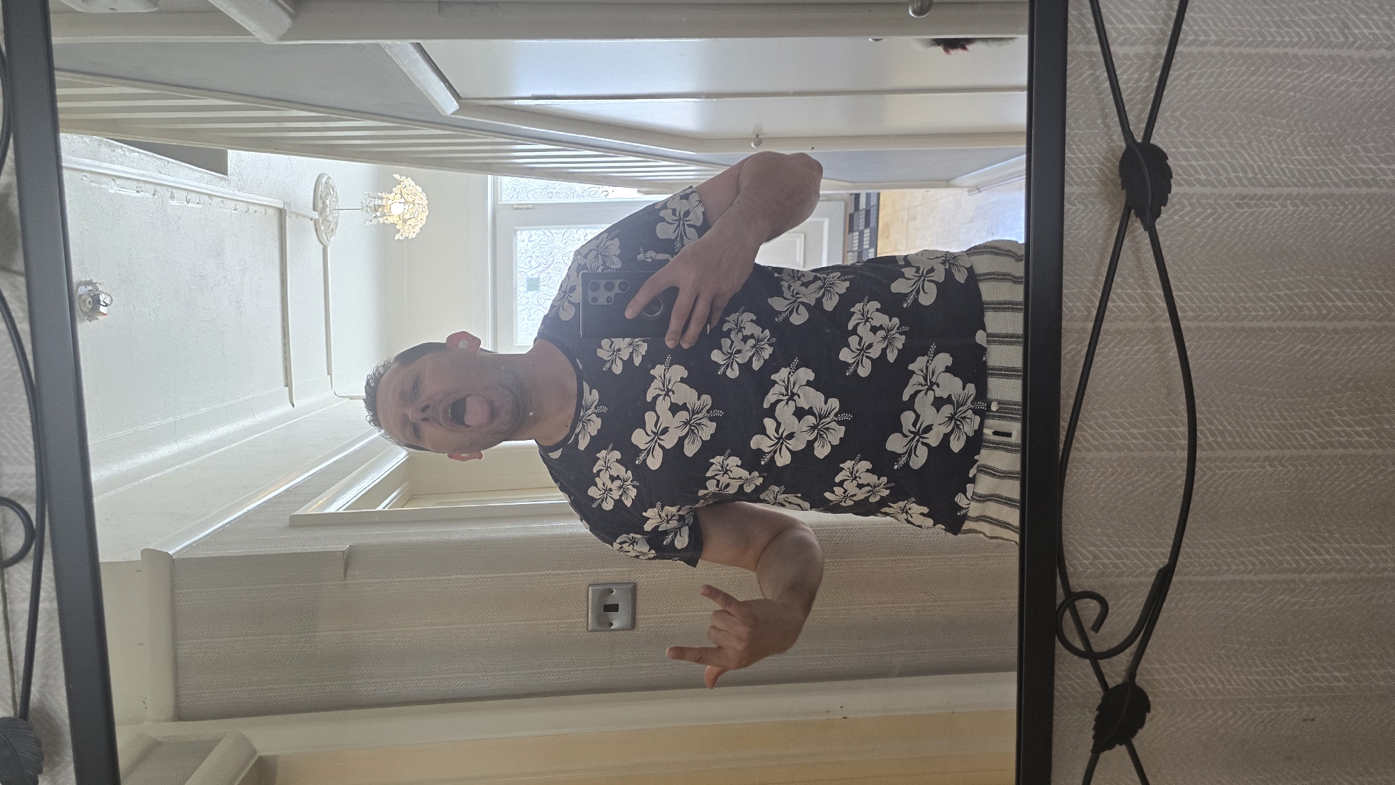 A middle-aged Caucasian man is reflected in a mirror, holding a smartphone in his left hand, taking a selfie. He sticks his tongue out and makes a rock-and-roll hand gesture with his right hand. He wears a dark t-shirt with a white floral pattern and striped shorts. The reflection shows a staircase in the background, suggesting the location is likely indoors, possibly a hallway. The mirror has a decorative black metal frame with leafy accents. The wallpaper has a subtle geometric pattern. The location in the city of London, United Kingdom, cannot be determined from the image.