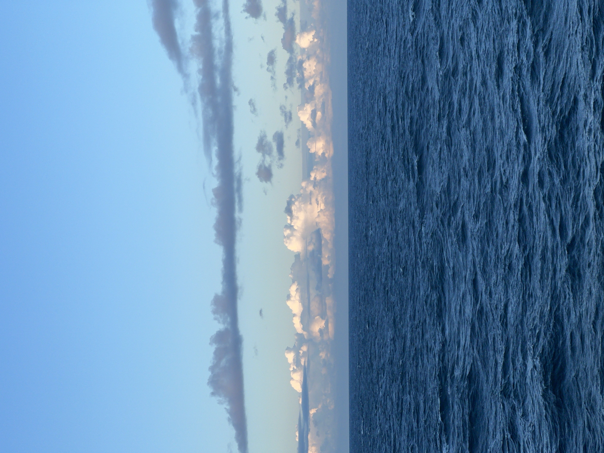 The image depicts a vast expanse of the ocean under a clear blue sky, with scattered clouds. The sea is a deep blue, with visible waves creating texture and movement across the surface. In the distance, a line of puffy white clouds is illuminated from below, suggesting the presence of sunlight near the horizon. Higher up, wispy, darker clouds drift across the sky. The scene is tranquil and natural, with no man-made structures or people visible. The location is not within a city, but rather an open body of water.