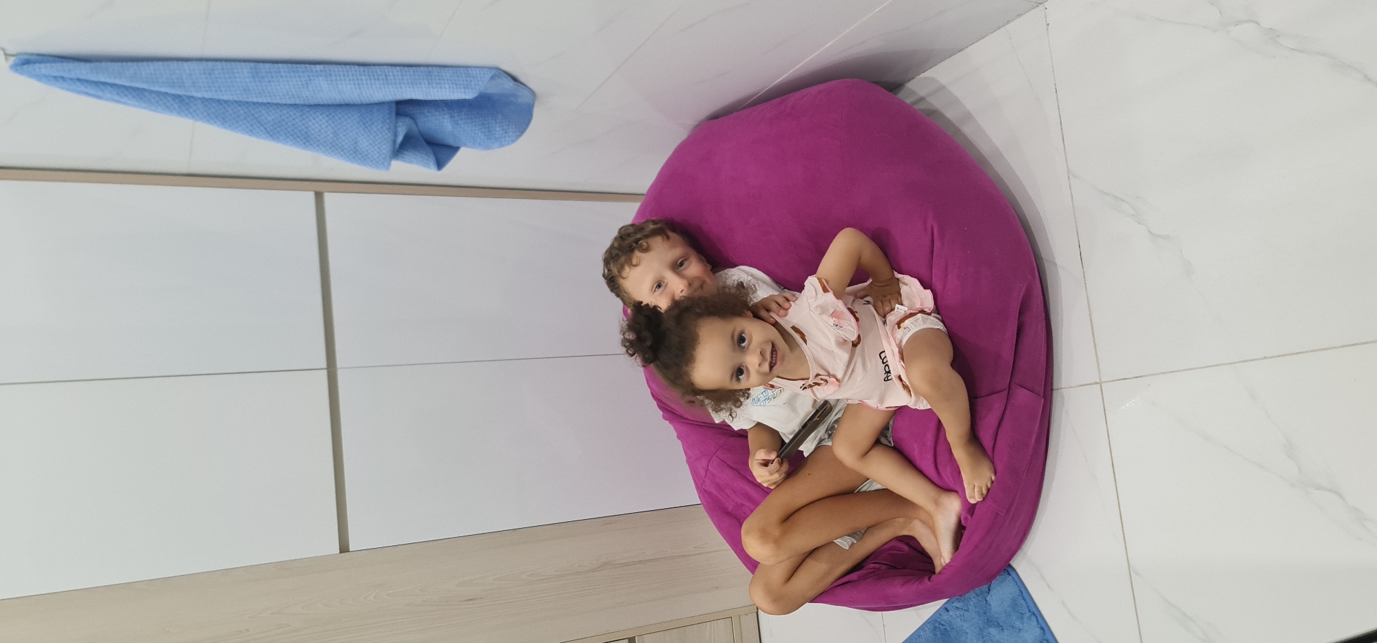 Two young children are sitting on a magenta beanbag chair in a room with white tile floors and walls. One child has curly brown hair and is wearing a light pink outfit. The other child has curly light brown hair and is wearing a white shirt. A light blue towel is hanging on the wall. The location is Đà Nẵng, Vietnam.