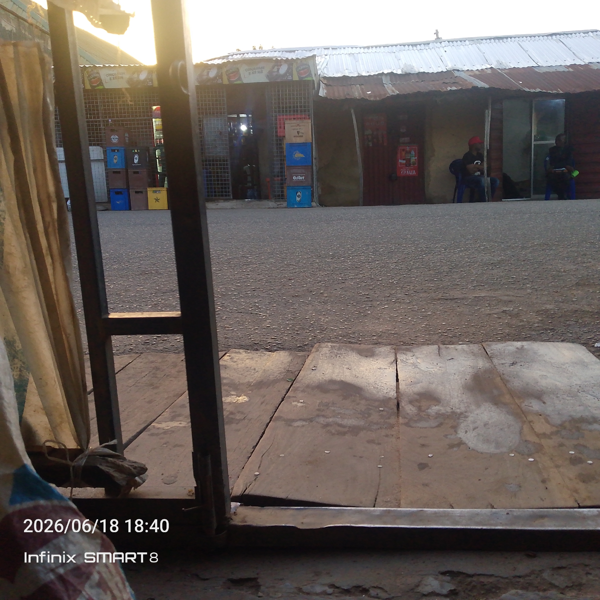 The image depicts a street-level view of a small outdoor market or shop with several stalls. In the foreground, a wooden platform is visible, with part of a metal gate and some draped fabric. The background shows a street with several crates of drinks stacked in front of the stalls. One stall has a metal grate door, and inside, some products are visible. Two men are seated on blue plastic chairs outside one of the stalls. The stall roofs are made of corrugated metal, some of which appear rusted. A sign with the text "75 NALIA" is visible on the side of one stall. The sky is bright, suggesting it is daytime. The timestamp in the lower left corner indicates the image was taken on June 18, 2026, at 18:40, and the device used was an Infinix SMART 8.