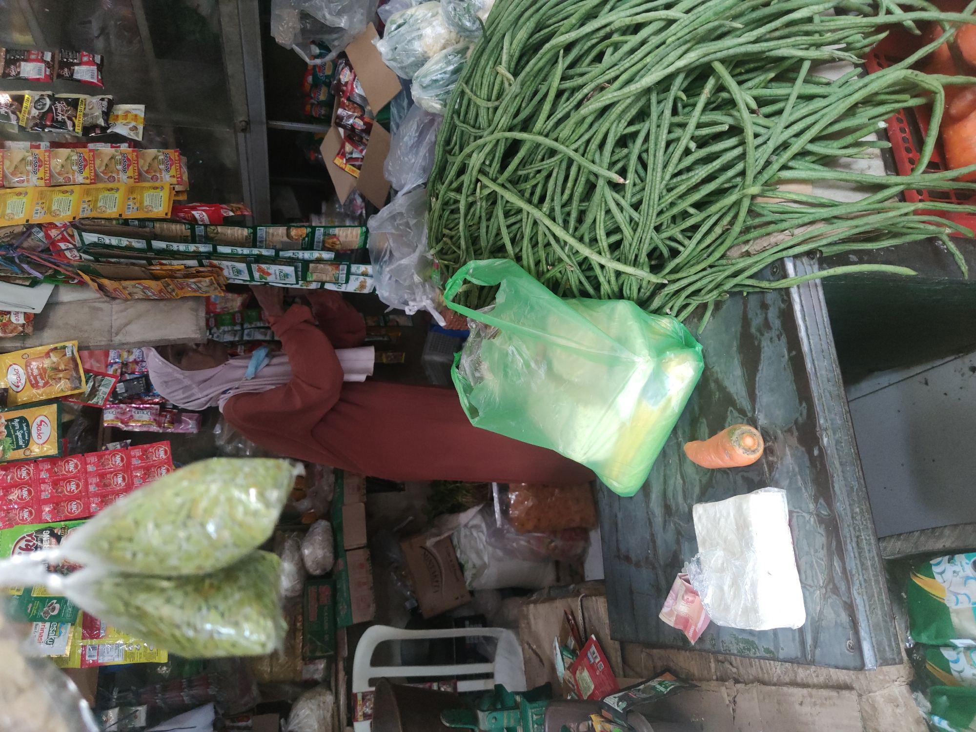 A person wearing a terracotta-colored dress and a light pink headscarf stands behind a table laden with produce and packaged goods in an outdoor market stall in Petiken, Indonesia. The person is facing to the right and appears to be interacting with items on the table or in the stall. In the foreground, a large pile of long green beans is spread across the right side of the table, with a green plastic bag partially obscuring some of them. To the left of the beans, a rectangular block wrapped in plastic, possibly tofu or tempeh, sits next to a single carrot. Behind the table, various packaged food items, including small sachets and larger bags, hang from racks or are stacked on shelves. A white plastic chair is visible to the far left, and a green weighing scale stands in front of it. The stall appears to be open-air or partially covered, with goods displayed both indoors and outdoors. The overall impression is of a bustling market scene with fresh produce and packaged food for sale.