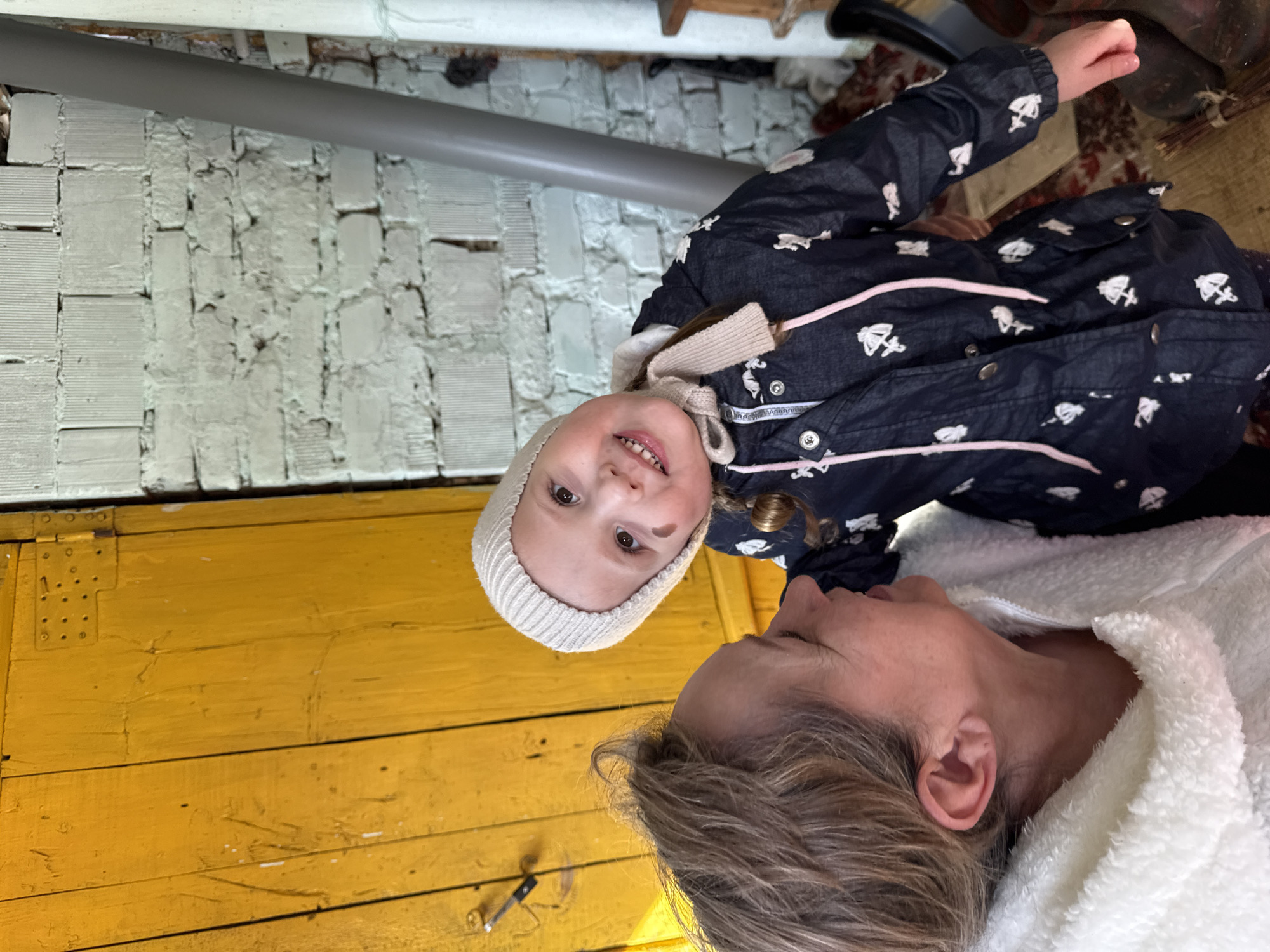 A photograph depicts a young girl and an older woman, seemingly a mother or grandmother, in an indoor setting. The girl is wearing a dark blue jacket with light-colored patterns, a light-colored hat, and appears to be smiling slightly. The woman is wearing a white fluffy robe or jacket. The background includes a yellow wooden wall and a white brick wall with a pipe running along it. The location is not discernible from the image.