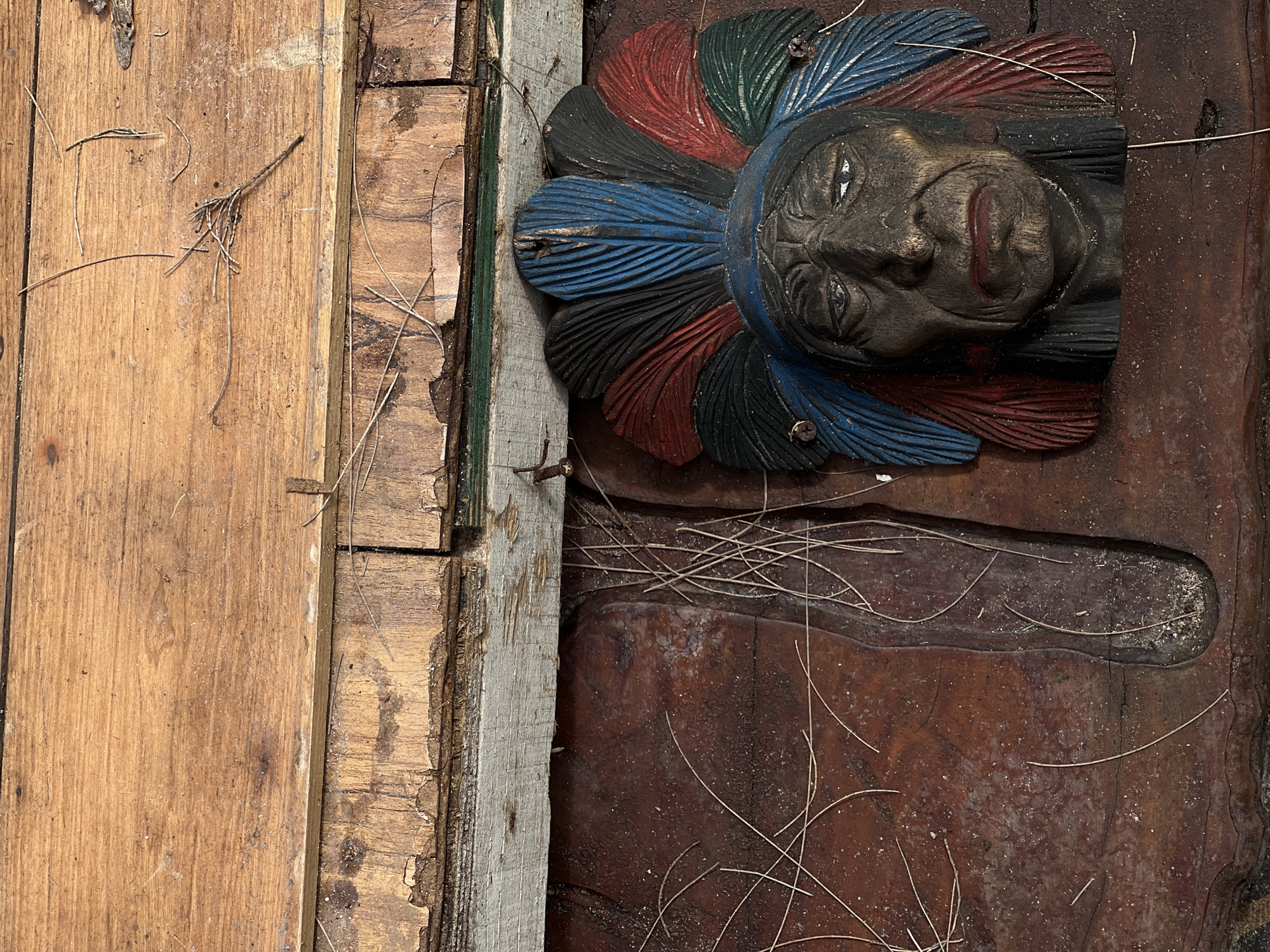An outdoor close-up shot captures a carved wooden sculpture of a Native American head. The sculpture is painted with vibrant colors, featuring a headdress with blue, red, black, and green feathers. The face has detailed wrinkles and painted eyes. The sculpture is mounted on a textured, dark brown surface. Above the sculpture, there are weathered wooden planks, with some appearing to be damaged and peeling. The scene is dusted with fine debris and thin, dry grass-like material. The location is described as Unknown, Unknown.