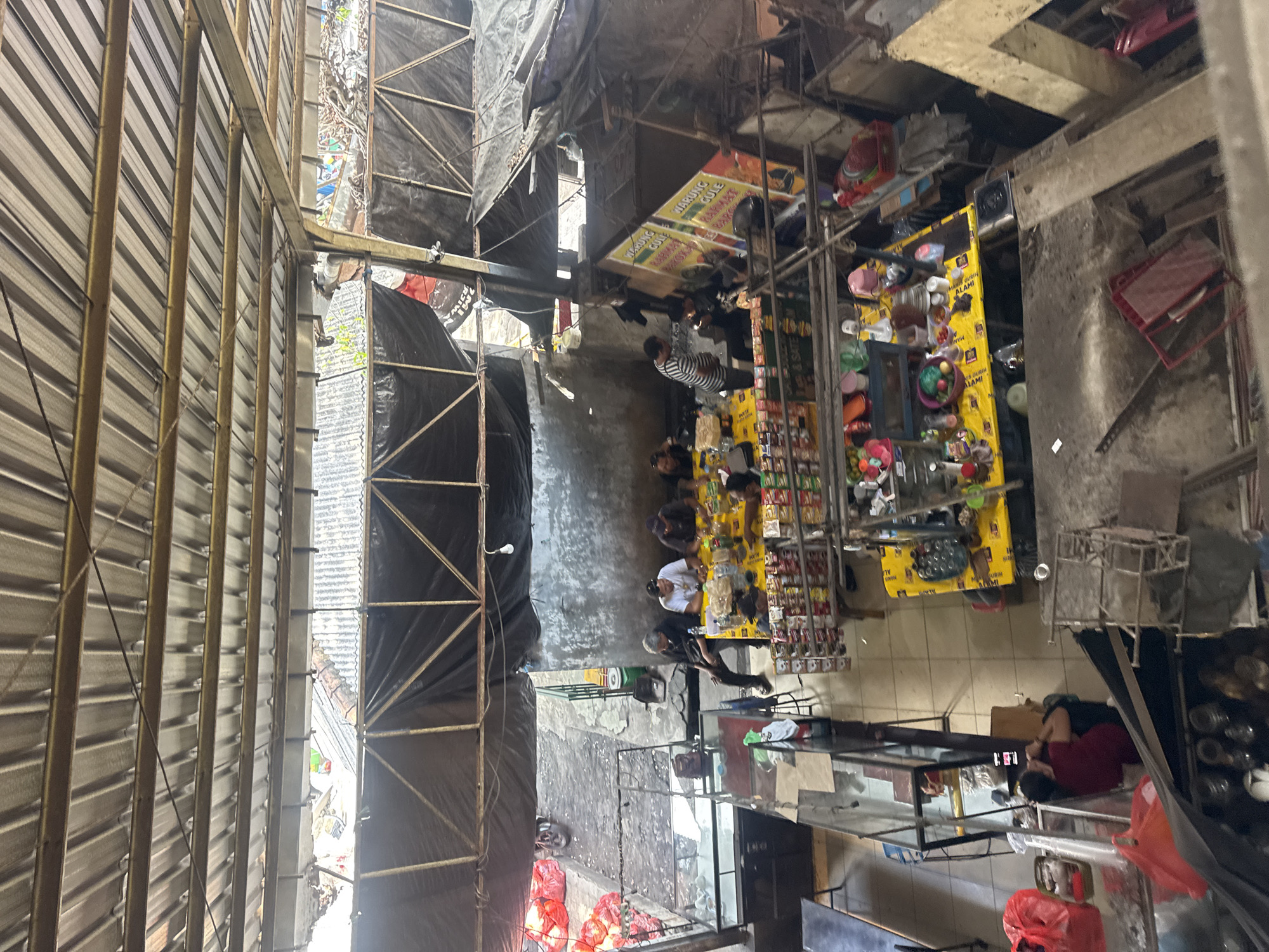 The image depicts an outdoor market scene, likely a food stall area, in Denpasar, Indonesia. The scene is viewed from an elevated perspective, looking down into an open-air structure covered by a corrugated metal roof supported by metal beams. Several individuals are seated around a yellow-covered table, suggesting a communal dining or gathering. They appear to be eating or conversing, with food and drinks on the table. In the background, more stalls are visible, some displaying goods for sale, including what appears to be packaged snacks and other products. Red plastic bags are stacked to the left of the frame, and a motorcycle is parked nearby. The environment is somewhat cluttered, with various materials and items scattered around the stalls, indicative of a bustling marketplace. A large banner advertising "WARUNG GULE" is prominently displayed on one of the stalls, further confirming the food stall aspect of the scene. The overall impression is that of a lively, informal gathering place within a local market in Denpasar.