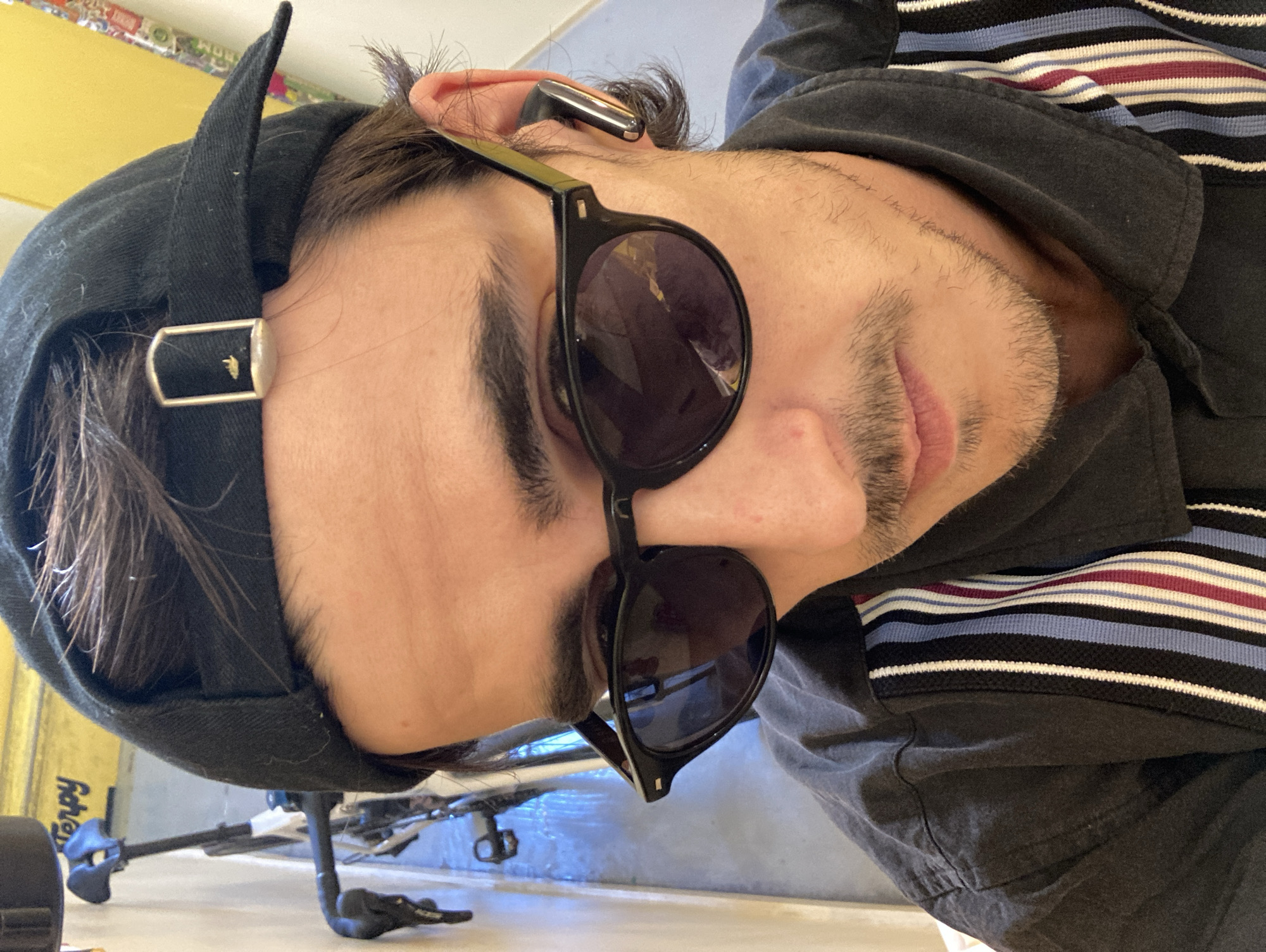 The image shows a close-up, medium shot of a young man wearing a black baseball cap, black sunglasses, and a dark collared shirt with colorful vertical stripes. He has a mustache and light stubble. A silver earbud is visible in his right ear. The background is blurred, but appears to be an indoor space with a bicycle in the lower left and a yellow wall to the right. The lighting suggests it is daytime. The man is looking towards the camera. The scene is not identifiable as being in Barcelona, Spain.