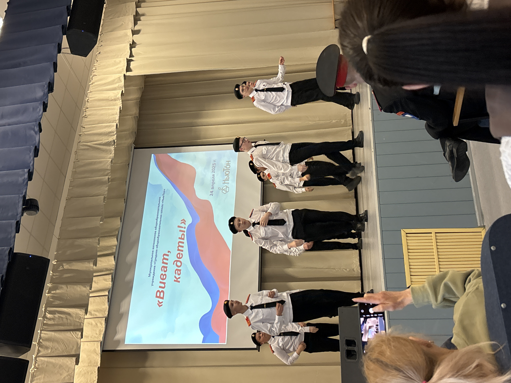 The image captures an indoor event in an auditorium or assembly hall, likely within a school setting in Chaikovsky, Russia. The scene depicts a group of young individuals, appearing to be cadets, performing or posing on a stage, dressed in white long-sleeved shirts, black ties, black pants, and black cadet hats or berets. They are arranged in synchronized positions, with one individual seemingly leading at the front, and others behind him.
Behind the performers, a large projector screen displays text and graphics. The visible text, as extracted by OCR, identifies the institution as "Муниципальное автономное общеобразовательное учреждение «Средняя общеобразовательная школа «Ньютон»" (Municipal autonomous general education institution "Secondary general education school "Newton"). The event title is "«Виват, кадеты!»" ("Vivat, Cadets!"), and the date "24 апреля 2025 г." (April 24, 2025) is prominently displayed, along with a logo for "Ньютон" (Newton). The screen features a background design of undulating red, white, and blue shapes.
In the foreground and to the sides, parts of an audience are visible, with individuals watching the performance. One person's hand is seen holding a smartphone, actively recording the event. The seating area consists of rows of folded blue and beige chairs. Large black audio speakers are suspended from the ceiling near the stage. The overall atmosphere suggests a formal school event or presentation, with the cadets showcasing their drill or performance skills.