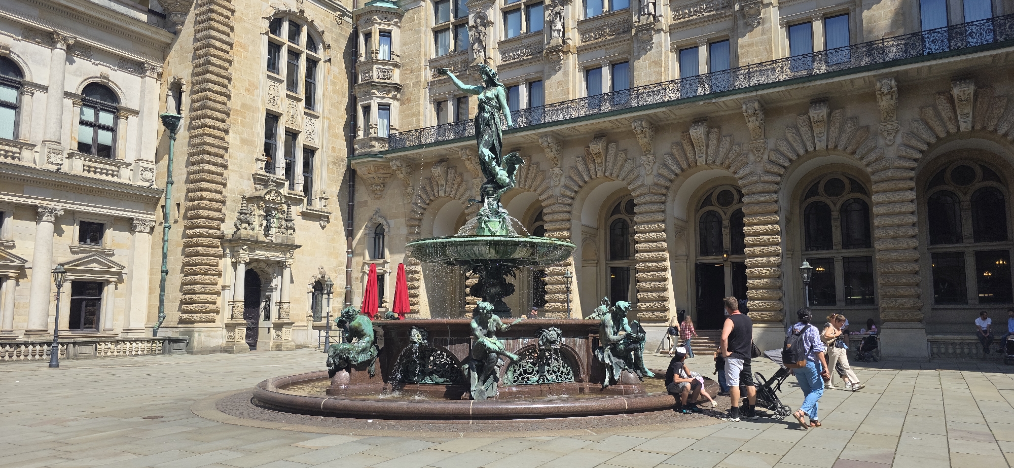 The image shows a daytime exterior view of a large, ornate building in Hamburg, Germany, likely the Rathaus (City Hall), given the architectural style and the prominent fountain in the foreground. The building is constructed of light-colored stone with numerous windows, balconies, and decorative architectural elements, including arches and columns. A large, multi-tiered bronze fountain with several sculptures of mythical figures and aquatic creatures is the central focus of the scene. Water is actively flowing from the fountain, creating a misty effect. The ground is paved with large, square stone tiles, forming a public square or courtyard. Several people are visible in the image, interacting with the fountain area and walking in the background. To the left of the fountain, two red umbrellas are set up. In the foreground, a few people are seated near the fountain, and others are walking or standing. A stroller is also present. The lighting suggests it is a sunny day with clear skies.