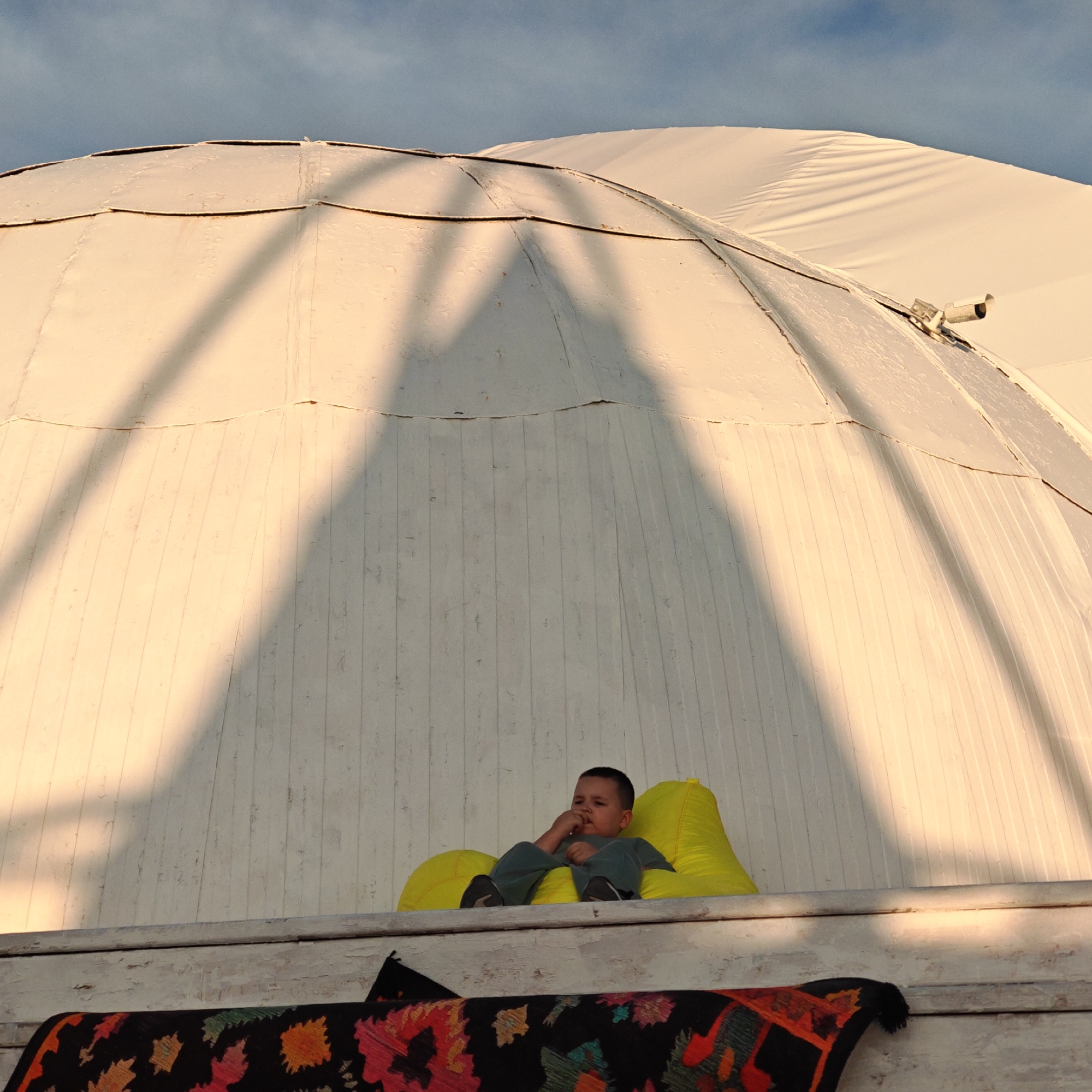 A low-angle shot captures a young boy sitting on a bright yellow beanbag chair, positioned on a white wooden ledge. The boy is looking down with his hand to his mouth, appearing to be in a pensive or thoughtful state. He is wearing a dark blue or gray t-shirt and pants. Behind him, the curved white surface of a domed structure dominates the background, casting long, angular shadows across its surface. The structure appears to be made of panels, with visible seams. The sky above is a clear, pale blue. A small, dark rug with colorful geometric patterns is visible on the ledge in front of the beanbag chair. The setting appears to be outdoors, possibly within a park or recreational area in the urban settlement of Shchyolkine, Ukraine. The lighting suggests it is either early morning or late afternoon, with the sun casting warm, directional light.