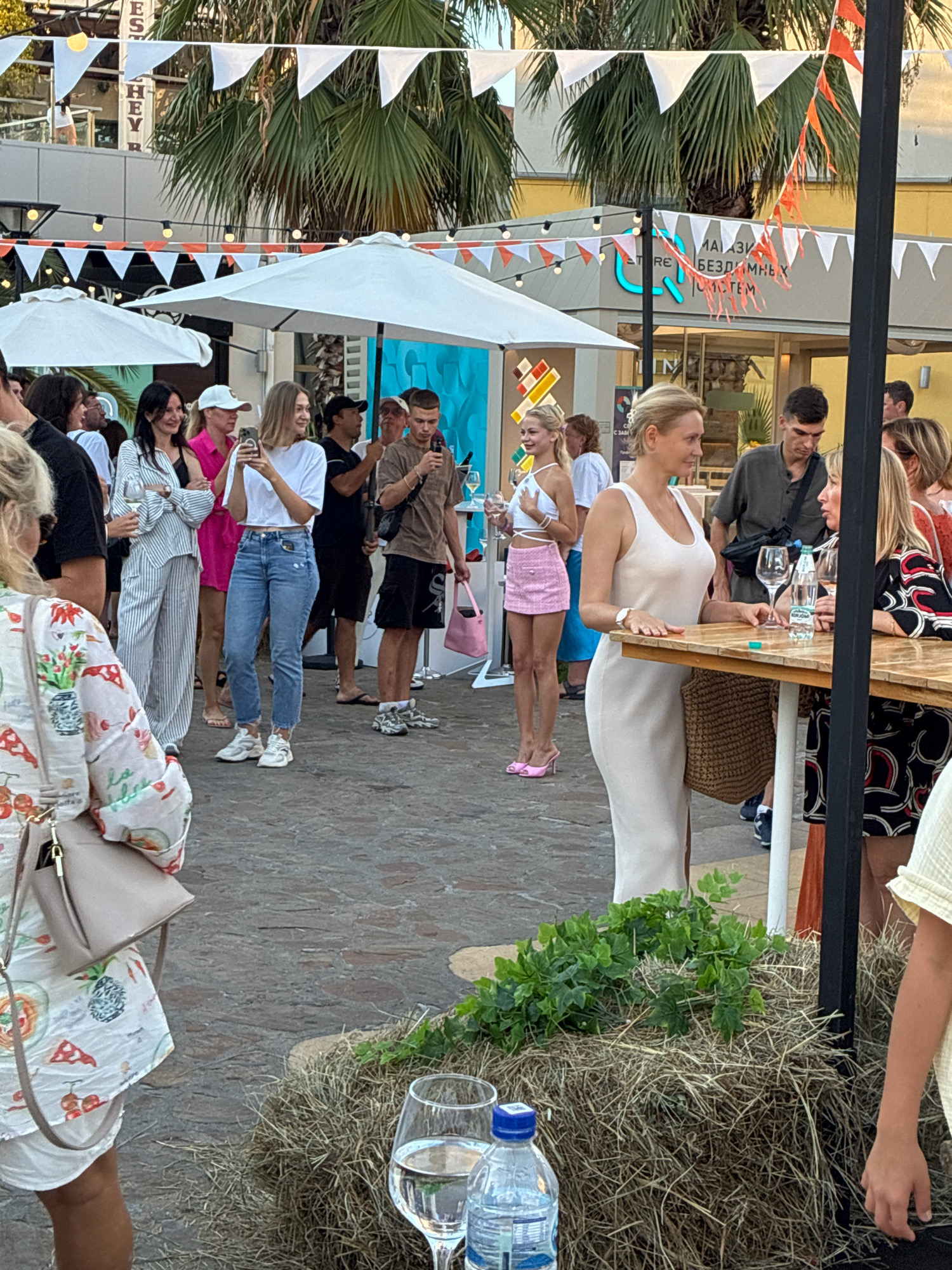 In Sochi, Russia, an outdoor event is underway, possibly a festival or market, characterized by decorative flags and white umbrellas providing shade. Several people are present, engaging in various activities such as talking, holding phones, and looking at their surroundings. In the foreground, a woman in a white dress stands near a table, while a person in a patterned dress is seated to her right. The environment includes a hay bale with greenery, a table with drinks, and a cobblestone ground. The background reveals buildings with signage, palm trees, and more attendees.