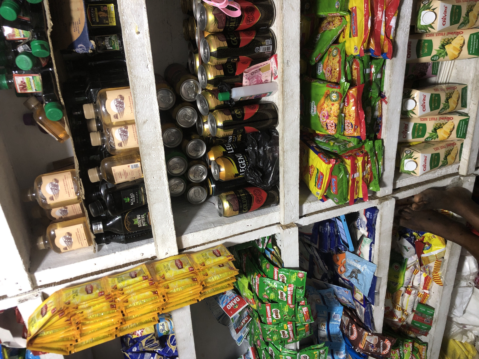 The image depicts the interior of a small convenience store, likely a kiosk or a corner shop, in Ado, Nigeria. The shelves are densely packed with various retail items, primarily food and beverages. Prominently displayed are numerous packets of Lipton yellow label tea hanging from the left side of the frame. Below these are several green and white packets of Milo beverage mix and other food items like Dano milk and Indomie instant noodles.
The shelves also contain an assortment of bottled drinks, including what appear to be alcoholic beverages in clear and dark glass bottles, some with green caps and labels that are difficult to read but suggest brands like "Kolawole Bitters" and other liquors. Several canned drinks, including "Goldberg" and "Legend" beer, are stacked on the shelves.
A bottle of hand sanitizer or a similar liquid product and Nigerian Naira currency are visible on a shelf alongside the canned drinks. On the bottom shelf, several cartons of Chivita Pineapple Juice are arranged.
In the lower right corner of the image, the feet and legs of a person are visible, suggesting they are either behind the counter or a customer seated very close to the merchandise. The environment appears somewhat cluttered, typical of a small, busy retail space. The lighting is uneven, with some areas well-lit and others in shadow, suggesting the use of artificial light. The shelves are made of wood and appear to be part of a makeshift shelving unit.