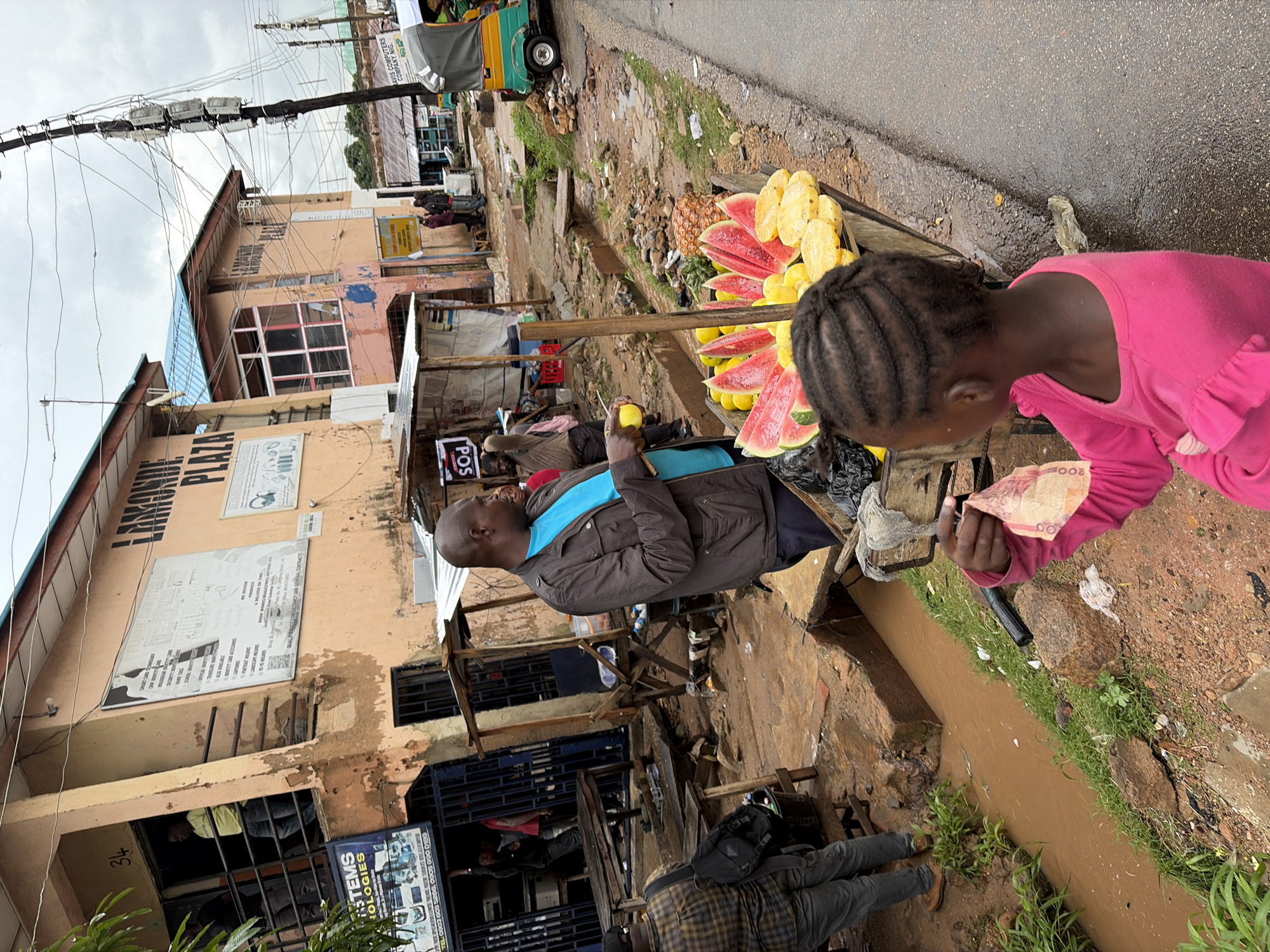 A busy street scene in Jos, Nigeria, features a fruit stand with a variety of fruits including watermelon and pineapple. A man in a brown jacket and blue shirt stands behind the fruit cart, holding a yellow fruit. A young girl in a pink shirt is in the foreground, holding a banknote. Several other people are visible in the background, as well as buildings and a three-wheeled vehicle. The street appears to be somewhat unkempt with a muddy ditch running alongside it.