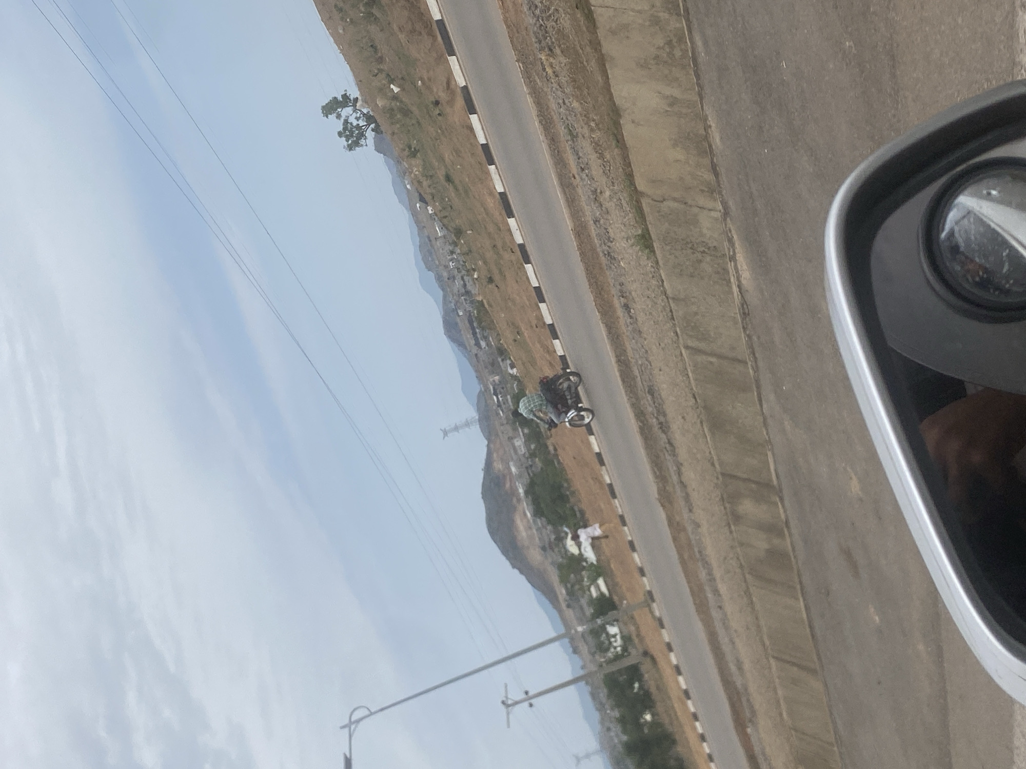 The image shows a roadside scene in what appears to be a hilly region outside of Abuja, Nigeria. A two-lane road, marked with black and white delineators, runs horizontally through the frame. Vegetation and settlements are visible along the hillside. A person is riding a motorcycle on the road. Utility poles and wires are visible against a cloudy sky. A car side mirror is in the foreground.