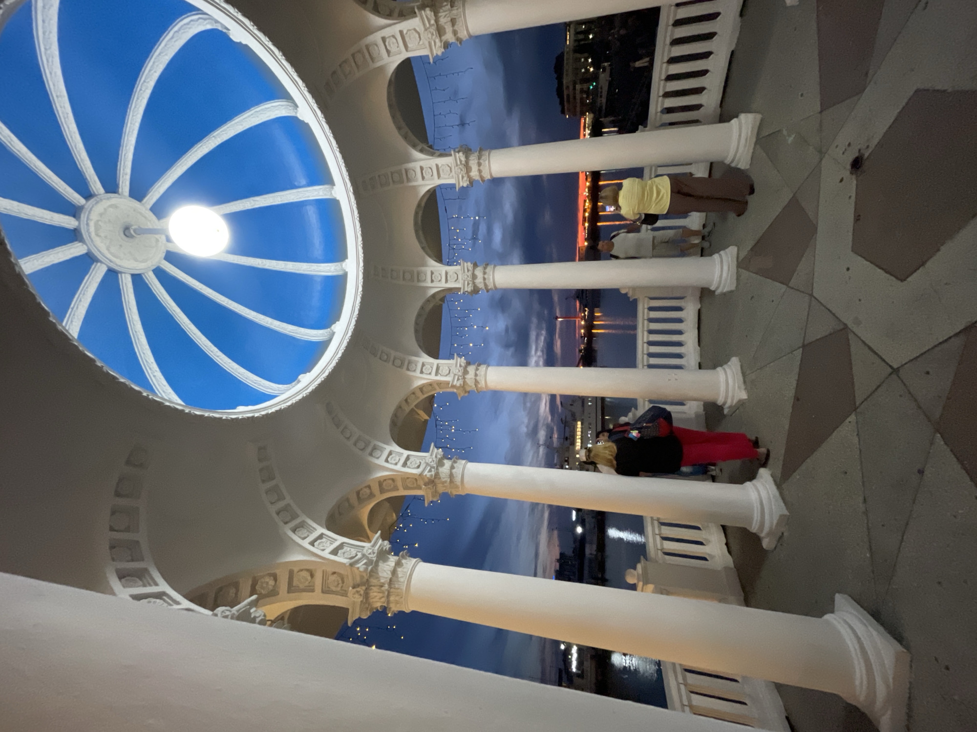 A wide-angle, eye-level shot captures the twilight view from an open-air rotunda with white Ionic columns and an ornate blue domed ceiling. The rotunda is decorated with hanging string lights. Four individuals are visible: two women and two men. One woman, wearing red pants and a black top, stands with her back to the camera, facing the water. Another woman in a yellow top and brown pants is also facing away, looking out at the scene. Two men, one in a white t-shirt and shorts, and another in a light-colored shirt and shorts, stand near the railing. They are all positioned on a paved area within the rotunda. Beyond the rotunda, a calm body of water reflects the lights of buildings and a ship docked in the distance. The sky is a blend of deep blue and dusky hues, indicating late evening. The architecture suggests a waterfront promenade, likely in Sochi, Russia, given the known presence of such structures along its coast, possibly near the central embankment or a park area.