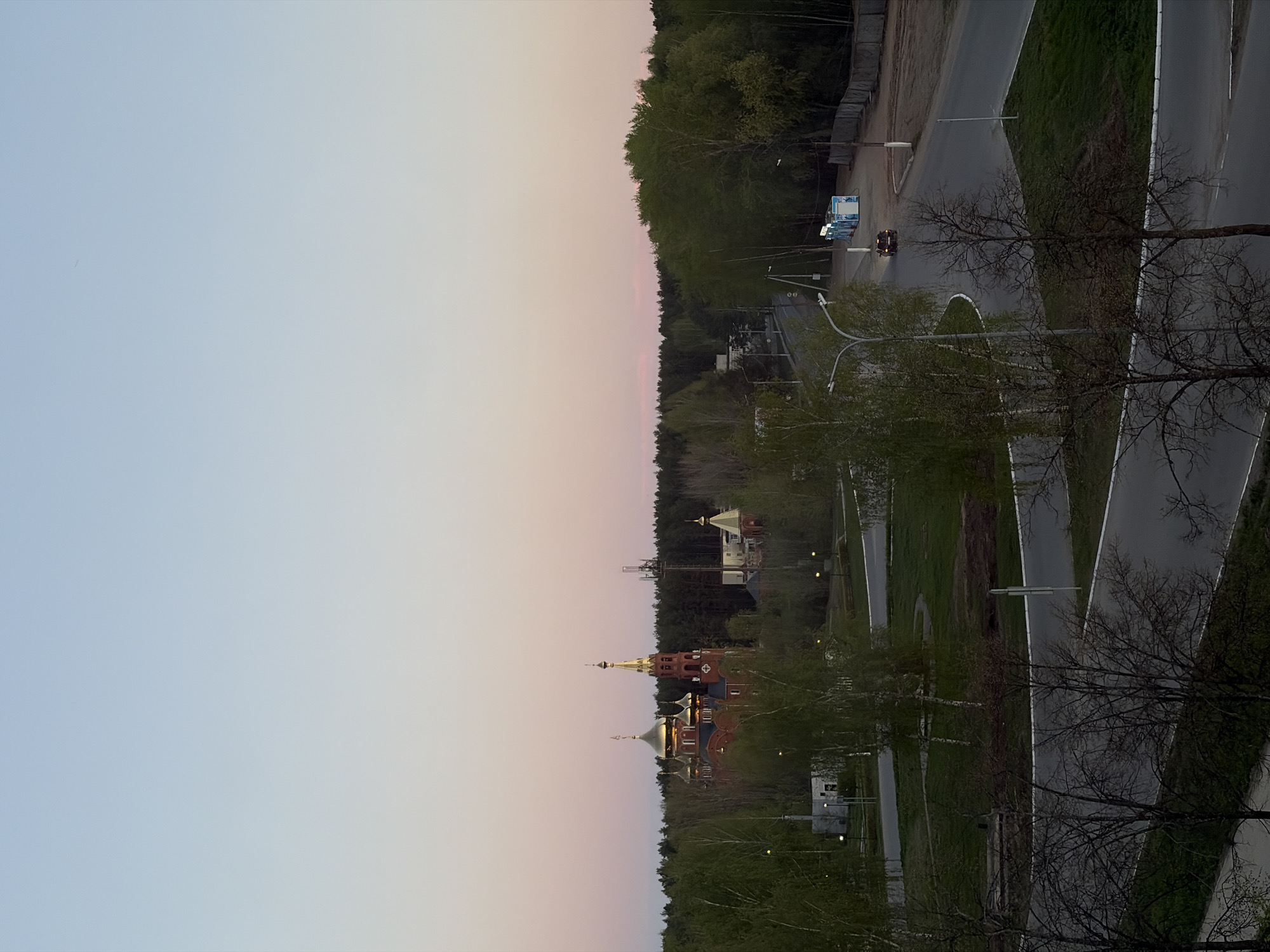 The image shows a landscape from an elevated perspective, likely a high-rise building, during either dawn or dusk. The sky transitions from a soft blue to pink.
The scene includes urban infrastructure and natural elements. There's a road network with multiple lanes and grassy medians. Several cars are visible on the road.
A prominent architectural feature is a Russian Orthodox church complex with golden domes and red brick buildings. The church is surrounded by green trees. Adjacent to the church, there is also another white building.
The scene is located in the city of Chaikovsky, Russia, as determined by the presence of the Russian Orthodox church complex.