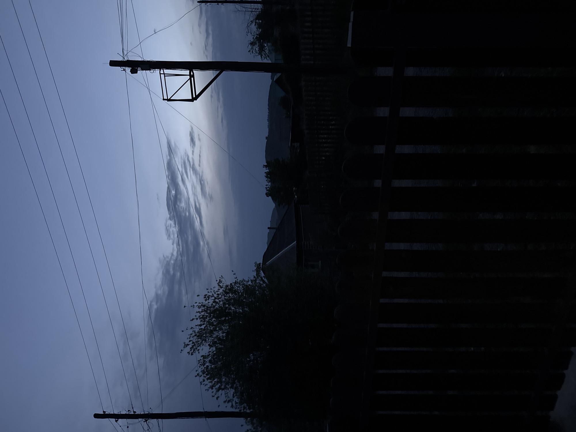 The image depicts a dark, twilight scene in the village of Soloneshnoye, Russia. A wooden fence, casting a deep shadow, dominates the foreground. Behind the fence, silhouetted against the dusky sky, are scattered houses with dark roofs, trees, and utility poles with numerous wires. The sky is a gradient of deep blue to lighter grey, with wispy clouds. The overall impression is one of quiet solitude in a rural setting at dusk.
