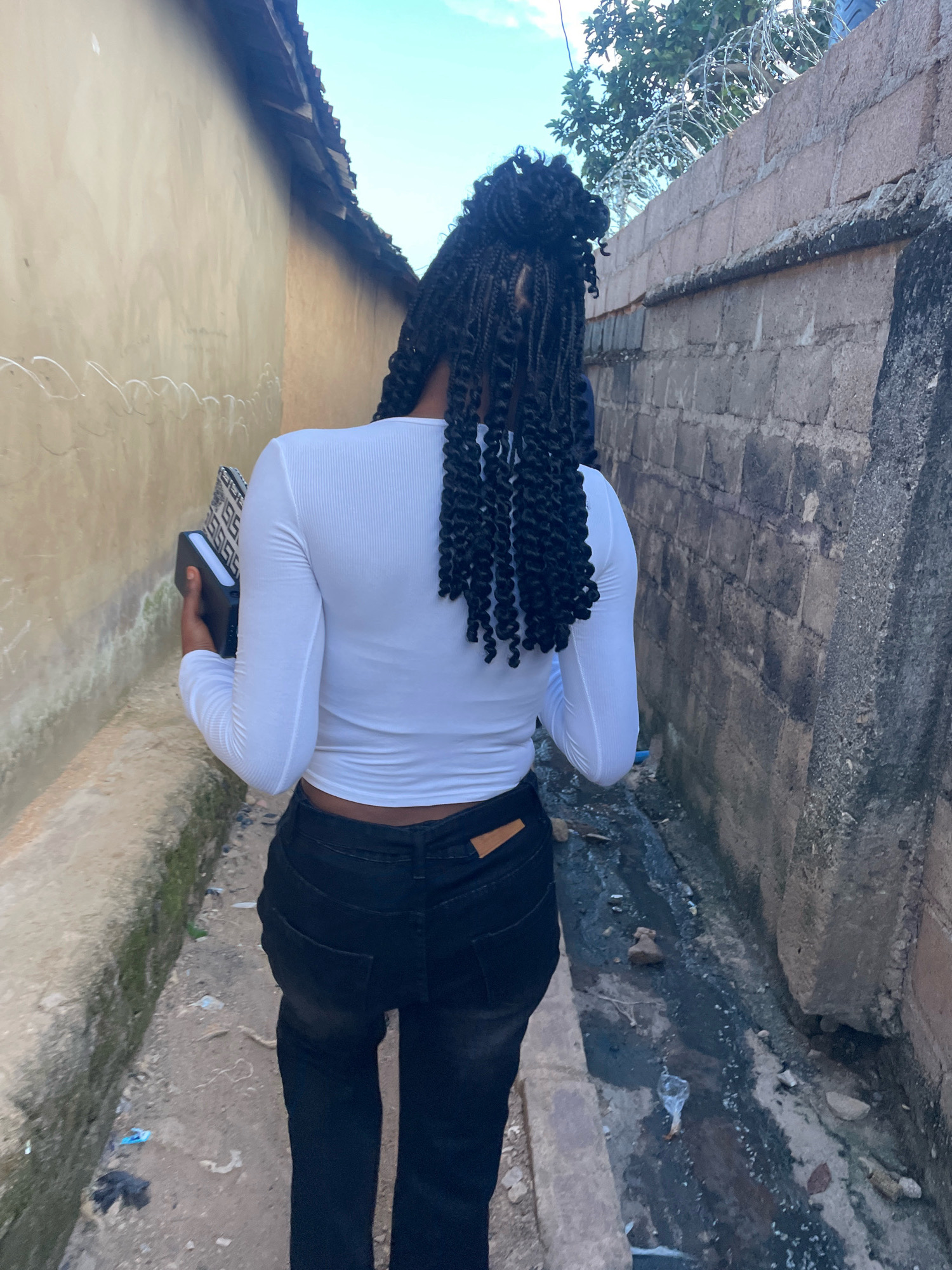 A person walks away from the camera down a narrow dirt alley. They are wearing a long-sleeved white crop top, black jeans, and have their hair in a braided style. The person is carrying a black and white patterned clutch or small bag. The alley is bordered by two walls: one a smooth, tan-colored wall on the left, and the other a rough, grey concrete block wall on the right topped with barbed wire. The ground of the alley is dirt and debris, with a small, dark, muddy drainage ditch running along the base of the concrete wall. Overhead, the sky is a pale blue with a few wispy clouds. The location is identified as Jos, Nigeria.