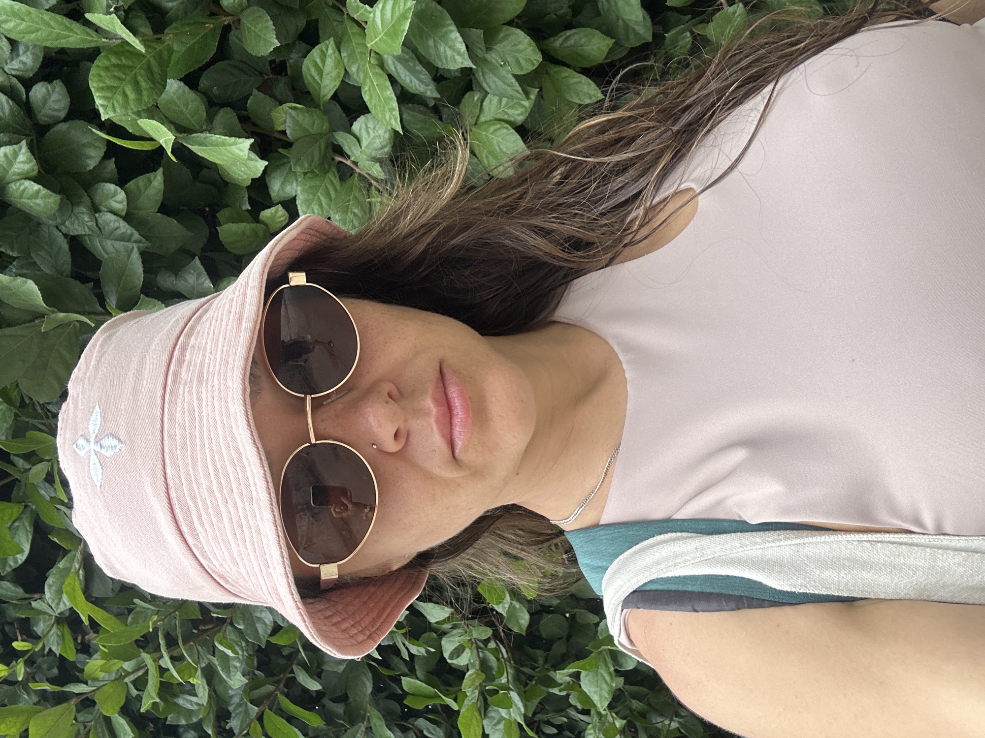A woman with brown hair and round sunglasses is wearing a pink bucket hat and a light pink shirt. She has a silver necklace and a nose piercing. She is lying down surrounded by green foliage. The image does not provide enough information to confirm the location in Bac Nha Trang Ward, Vietnam.