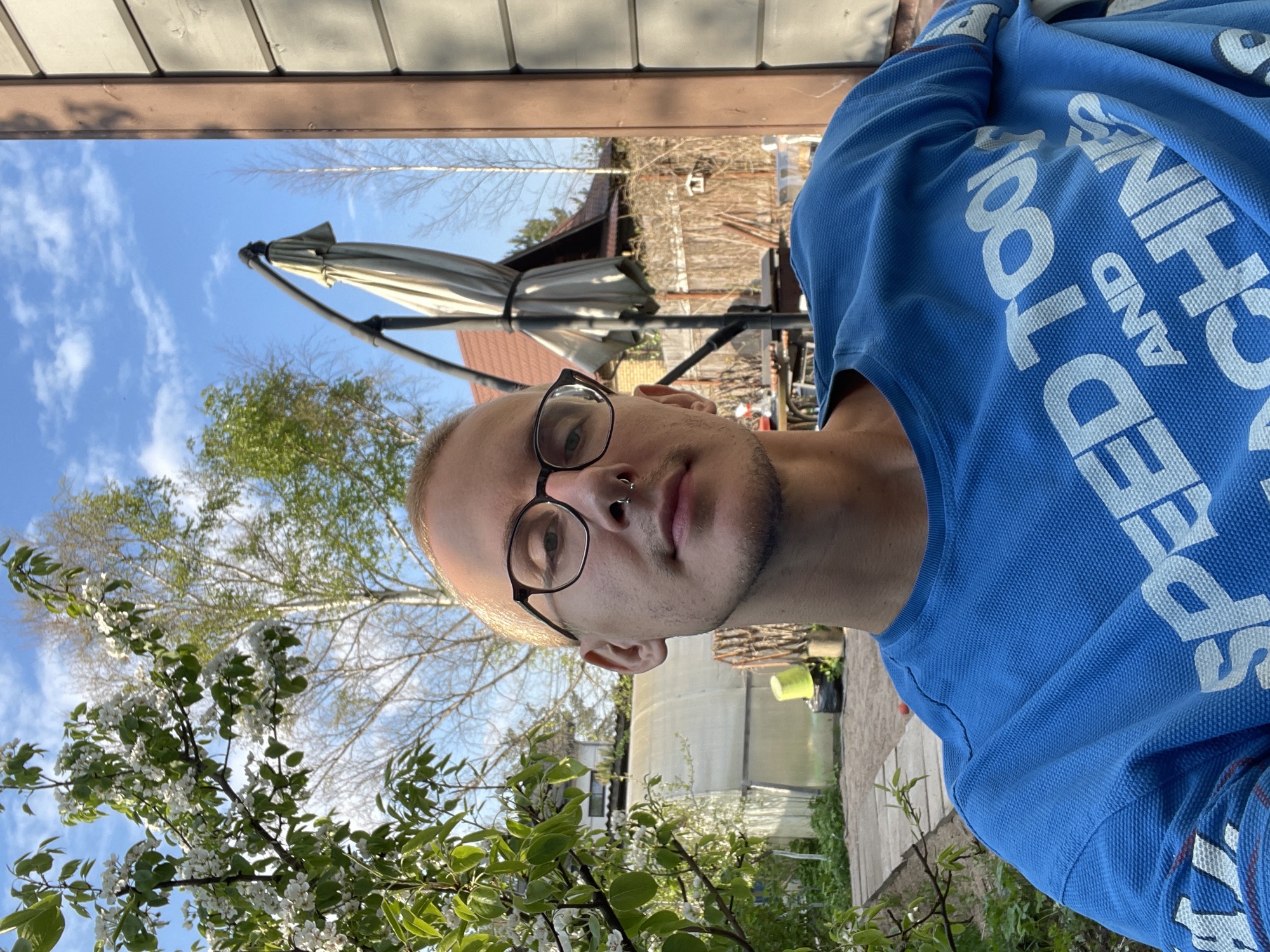 A medium-shot, slightly underexposed photograph taken outdoors in Sestroretsk, Russia shows a person wearing glasses and a blue shirt standing in a yard. The person is facing the camera. The background includes trees in bloom, a house, an umbrella, and a greenhouse. The sky is blue with scattered clouds. The image is slightly rotated counter-clockwise.