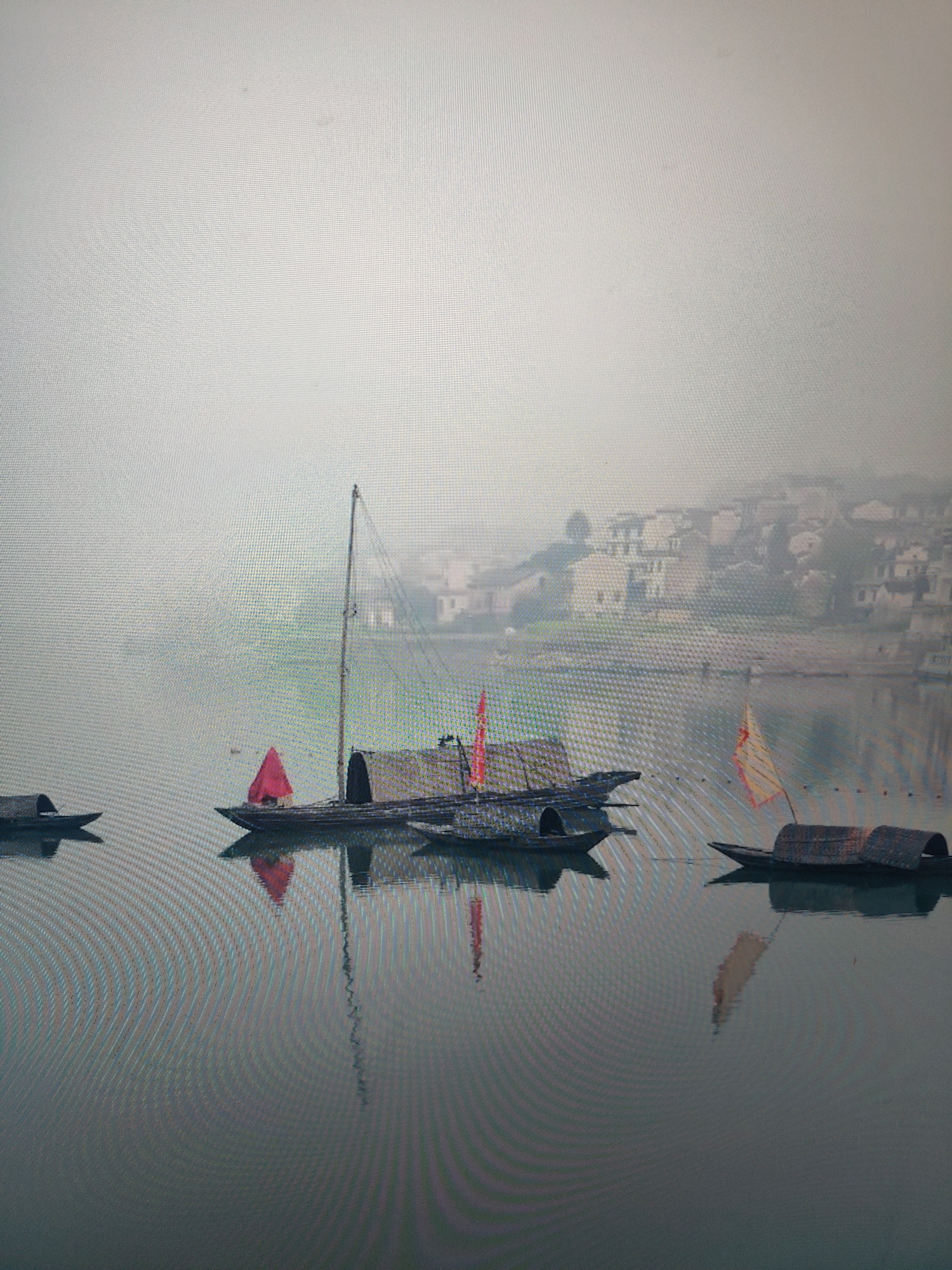 The image depicts several small boats on a body of water, likely a river or lake, on a foggy or misty day. The boats are traditional in style, with enclosed or covered sections and at least one with a mast and sail. Each boat is in a different location on the water, and the houses on the shore are difficult to make out due to the fog. The water appears calm with some visible ripples. The city is Unknown, Unknown.