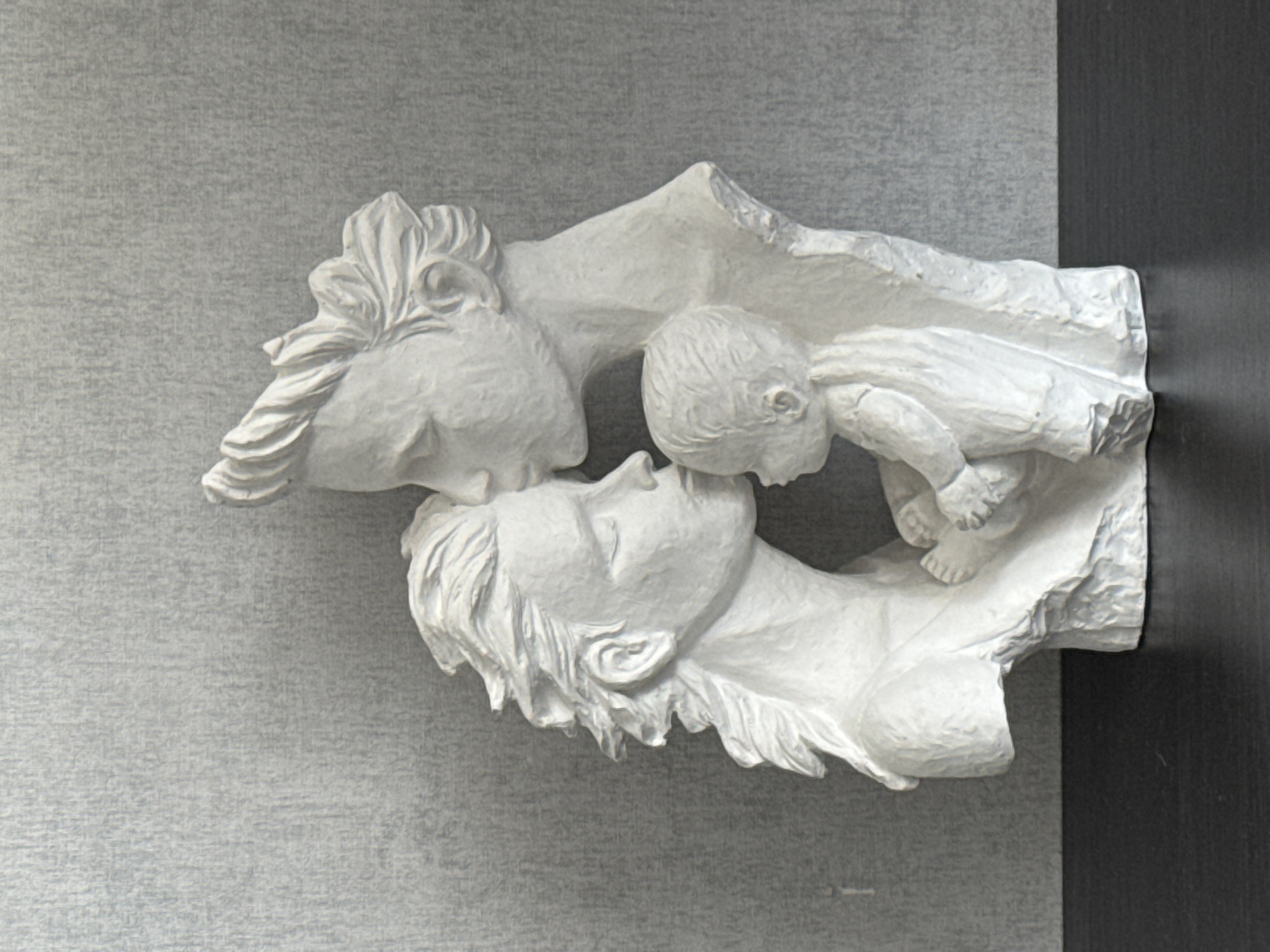 A white plaster sculpture of a family is displayed against a textured gray wall. The sculpture depicts a man kissing the forehead of a woman, who is leaning in to kiss a baby held in her arms. The figures are rendered in a stylized, minimalist manner. The sculpture rests on a dark, flat surface, likely a table or shelf. The background is a plain, textured wall. There is no indication of the sculpture's location in Cheboksary, Russia; this information is not present in the image.