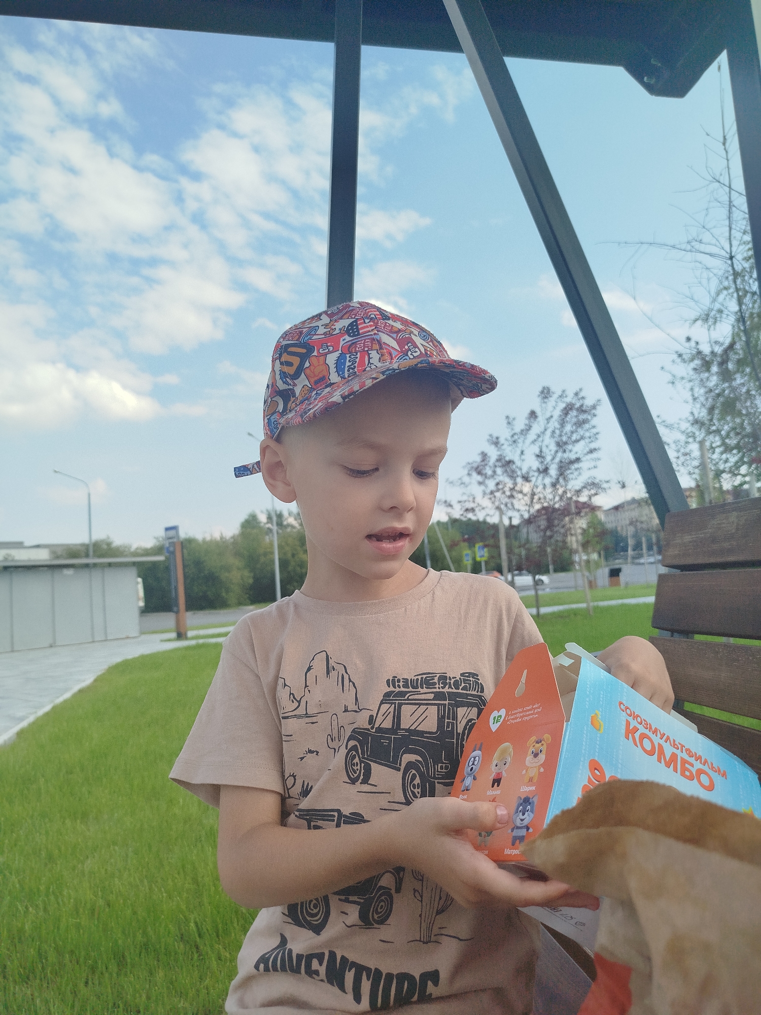 The image shows a young boy wearing a cap and a t-shirt with a desert-themed graphic, sitting on a wooden bench in an outdoor public space, possibly a park or a recreational area in Tyumen, Russia. The boy is holding a McDonald's Happy Meal box. The box has cartoon characters and text in Russian, including "Союзмультфильм КОМБО," indicating it's a special promotion with characters from Russian animated films. In the background, there is green grass, trees, buildings, and a cloudy sky. The overall setting appears to be a sunny day.